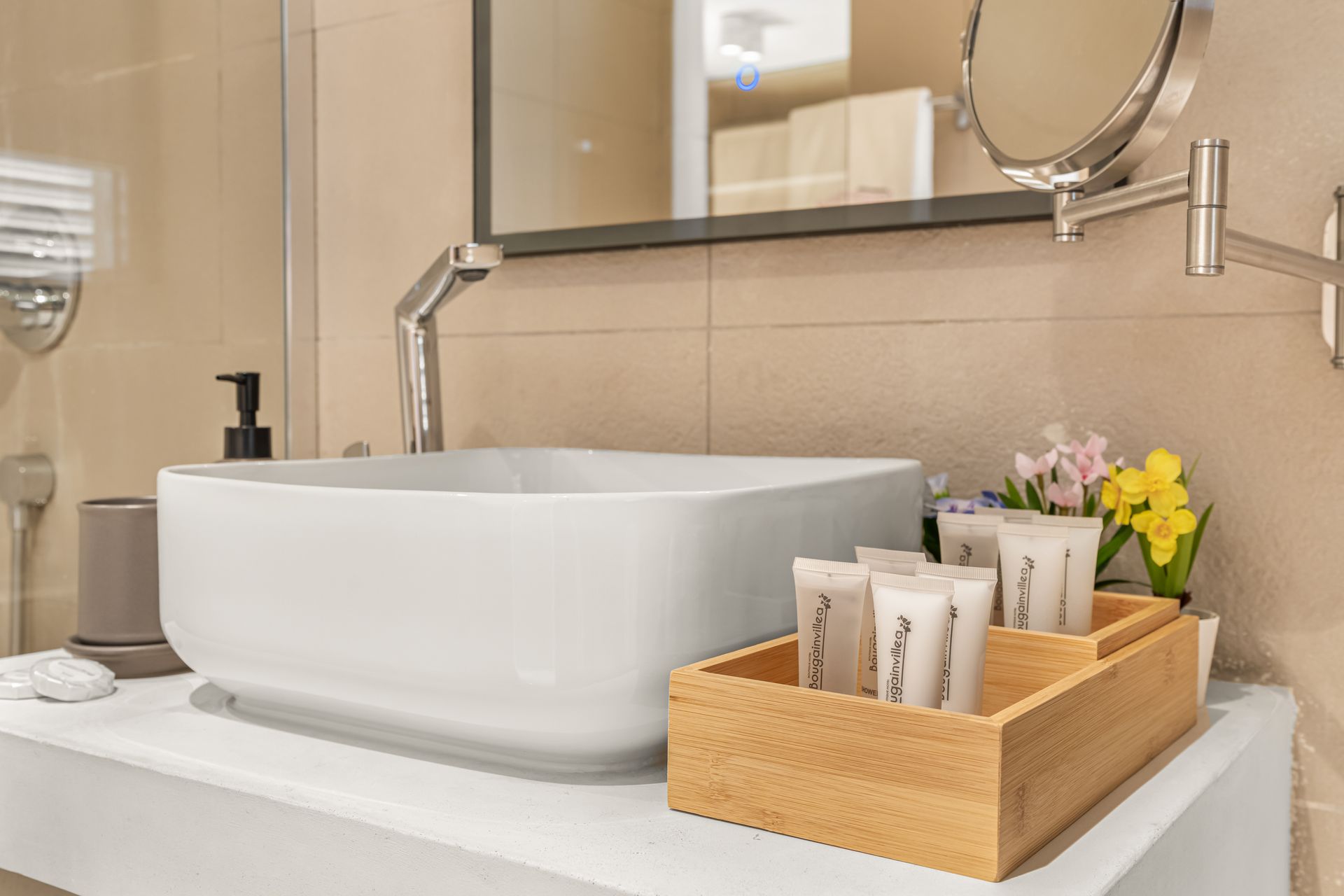 White bathroom sink with soap dispenser, toiletries in wooden tray, mirror, and flowers.