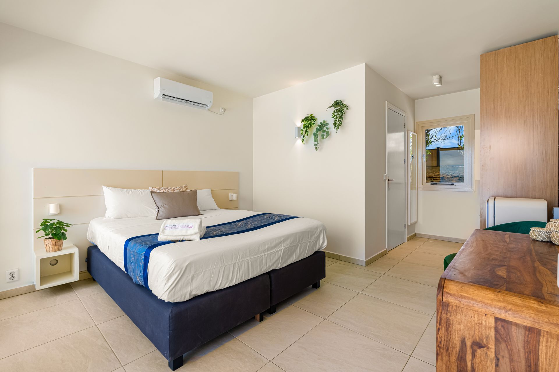 Bright bedroom with a neatly made bed, white walls, tiled floor, and a wooden desk by the window.