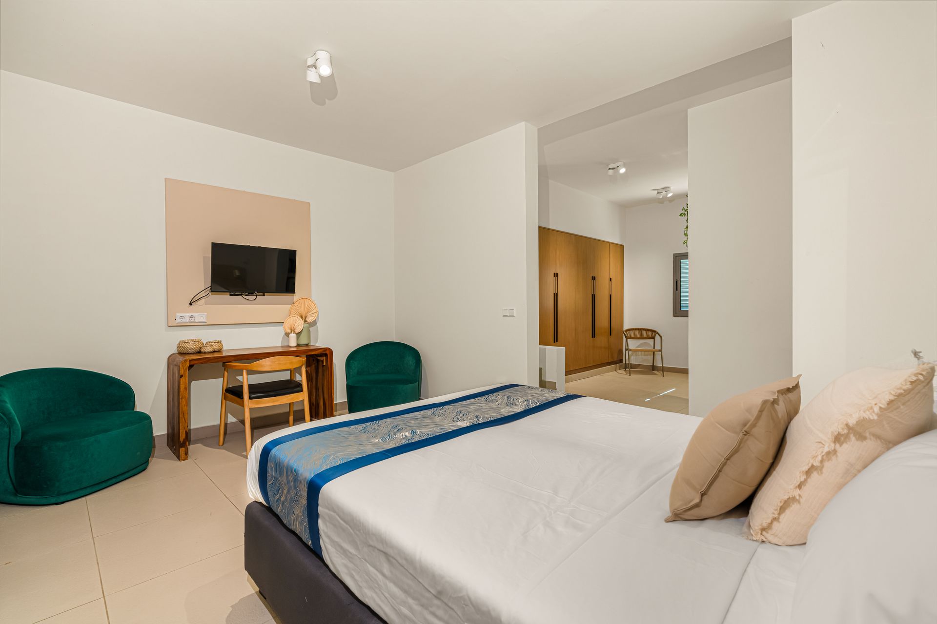 Bedroom with bed, desk, chairs, TV, and large doorway to another room. Light colors and modern furniture.