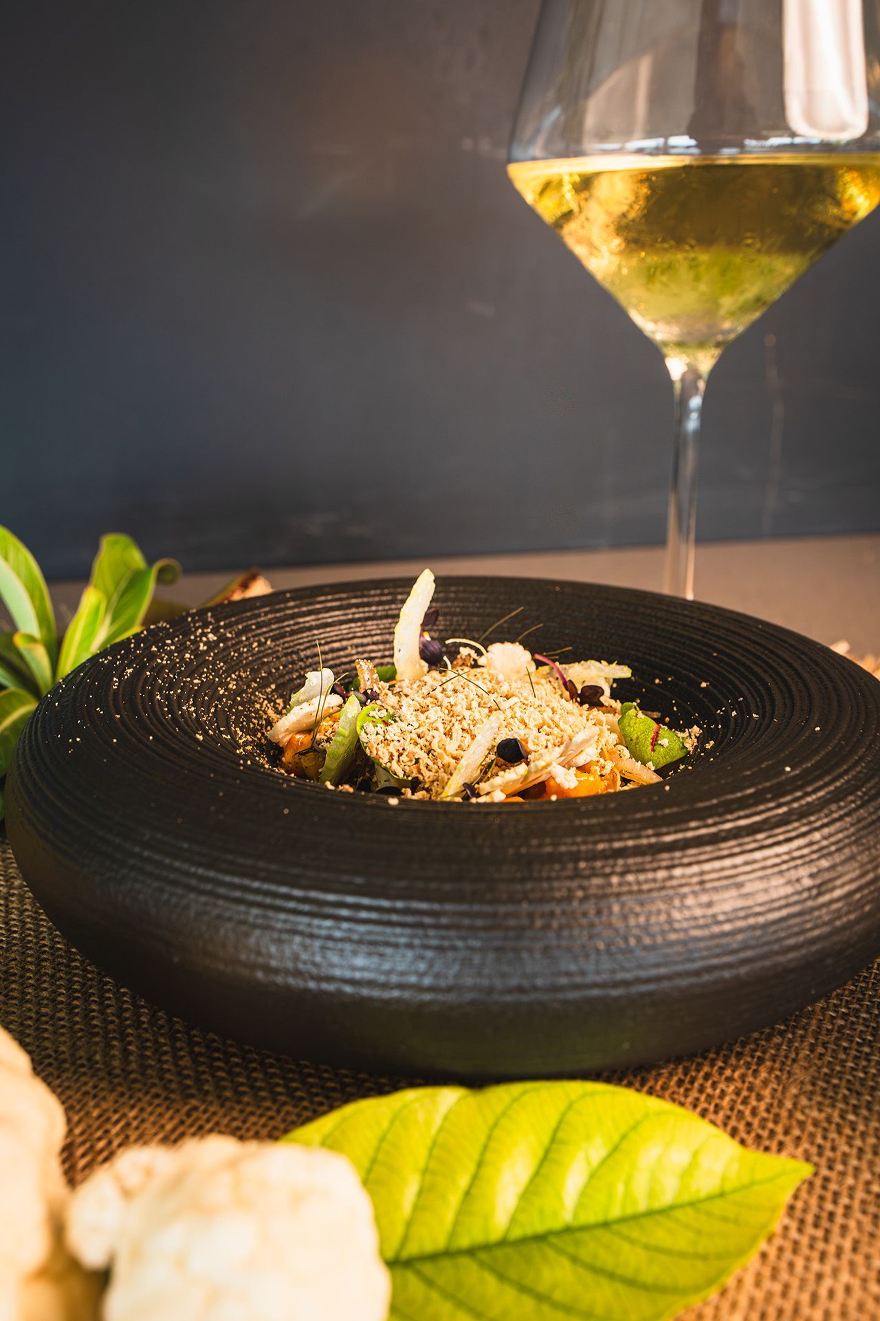 Black bowl of food topped with sesame seeds, next to a wine glass and cauliflower.