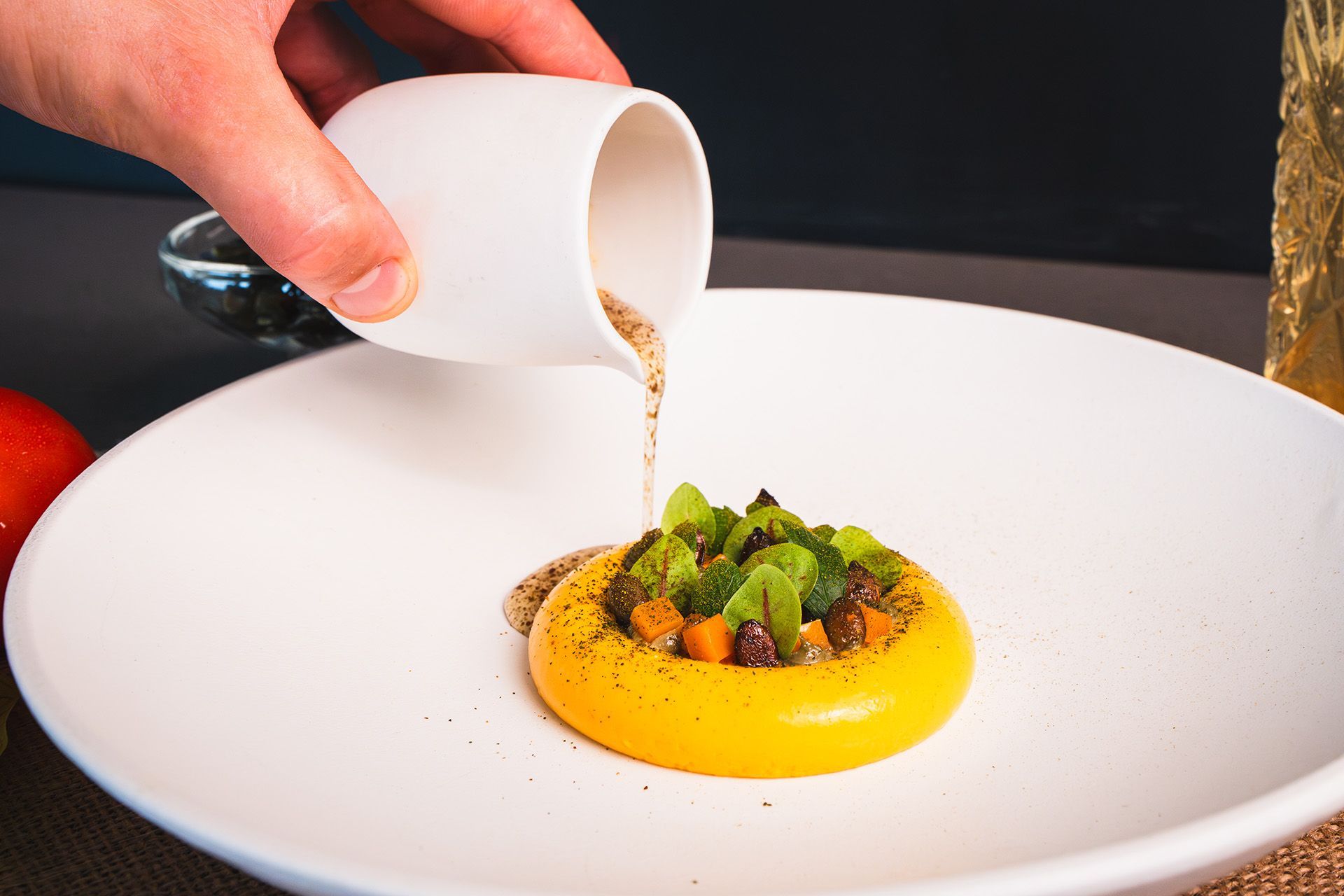 A hand pouring sauce over a yellow, circular food arrangement on a white plate.