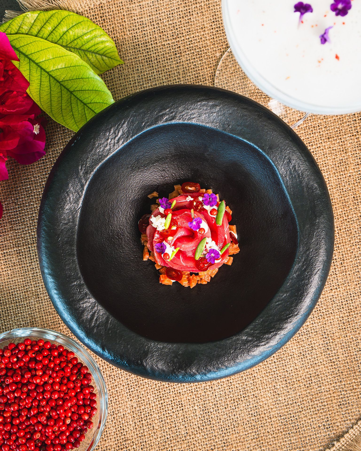 Black bowl with colorful food. Garnished dish, red berries, and floral elements on textured surface.