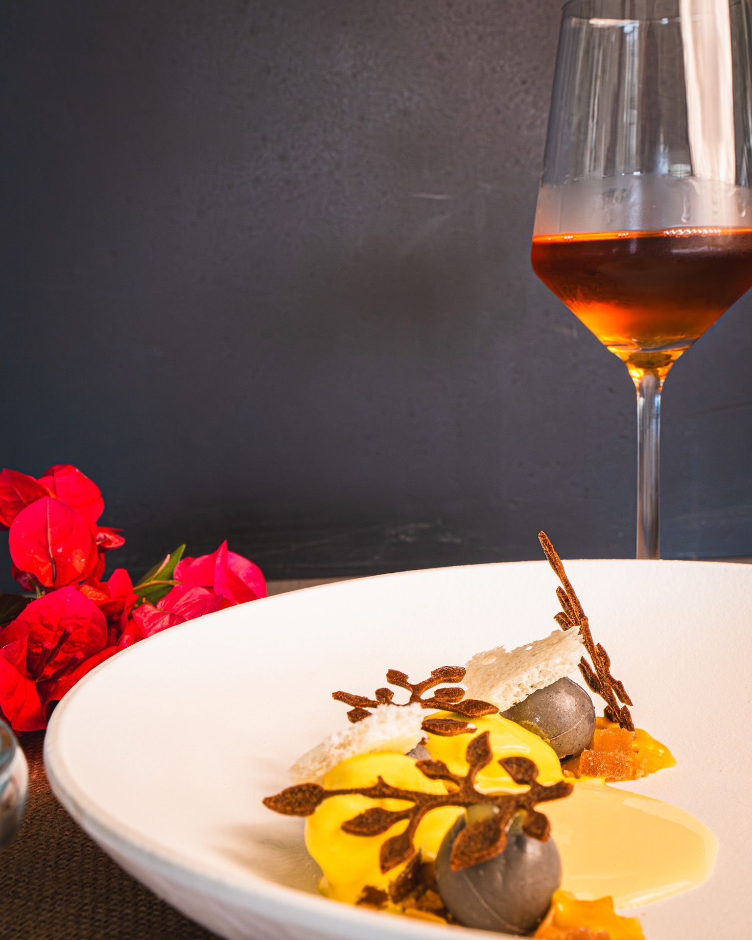 Dessert plate with yellow and gray elements, chocolate garnish, and glass of amber liquid.