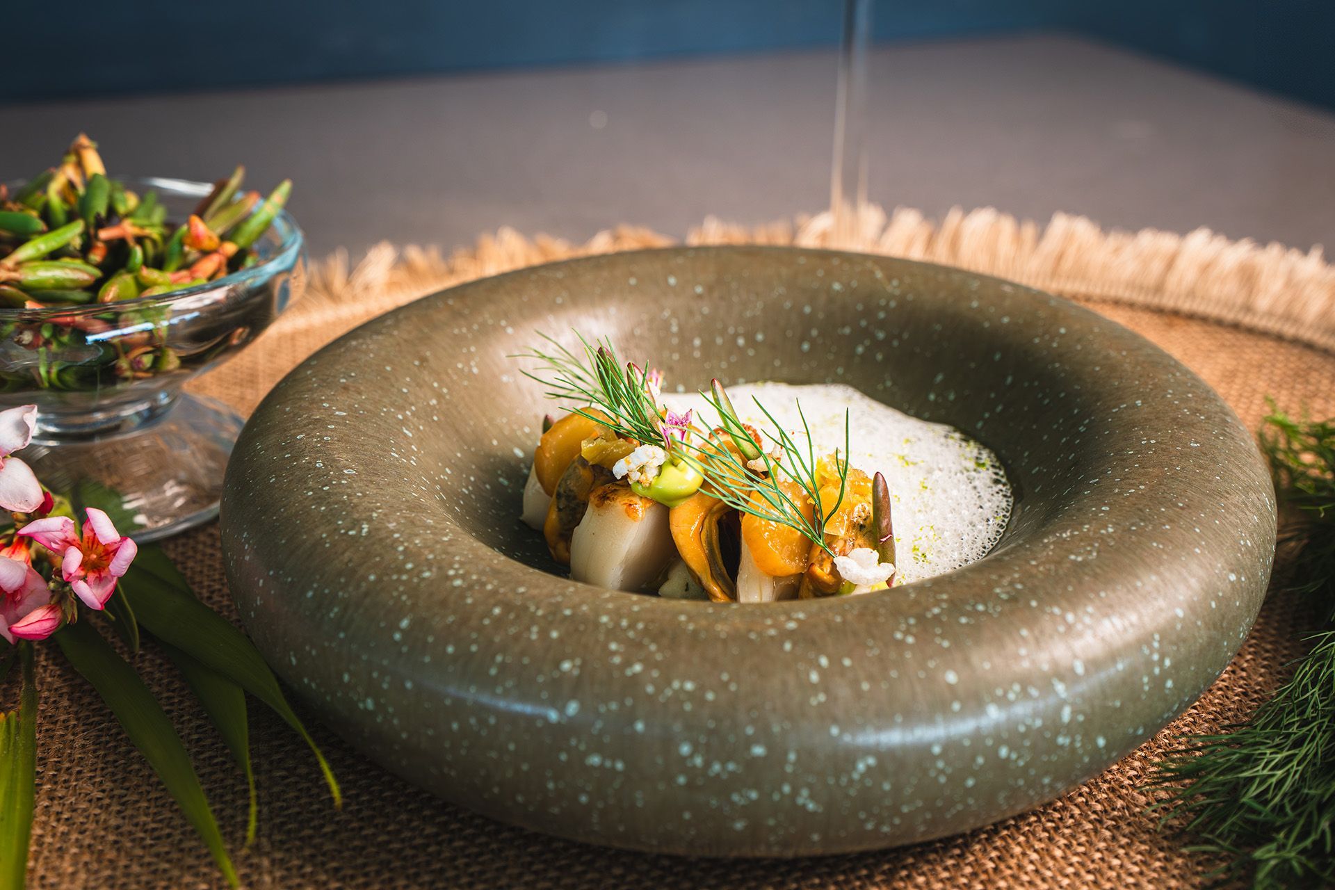 Close-up of a seafood dish in a circular gray bowl, garnished with herbs. A side dish is visible in a glass bowl.