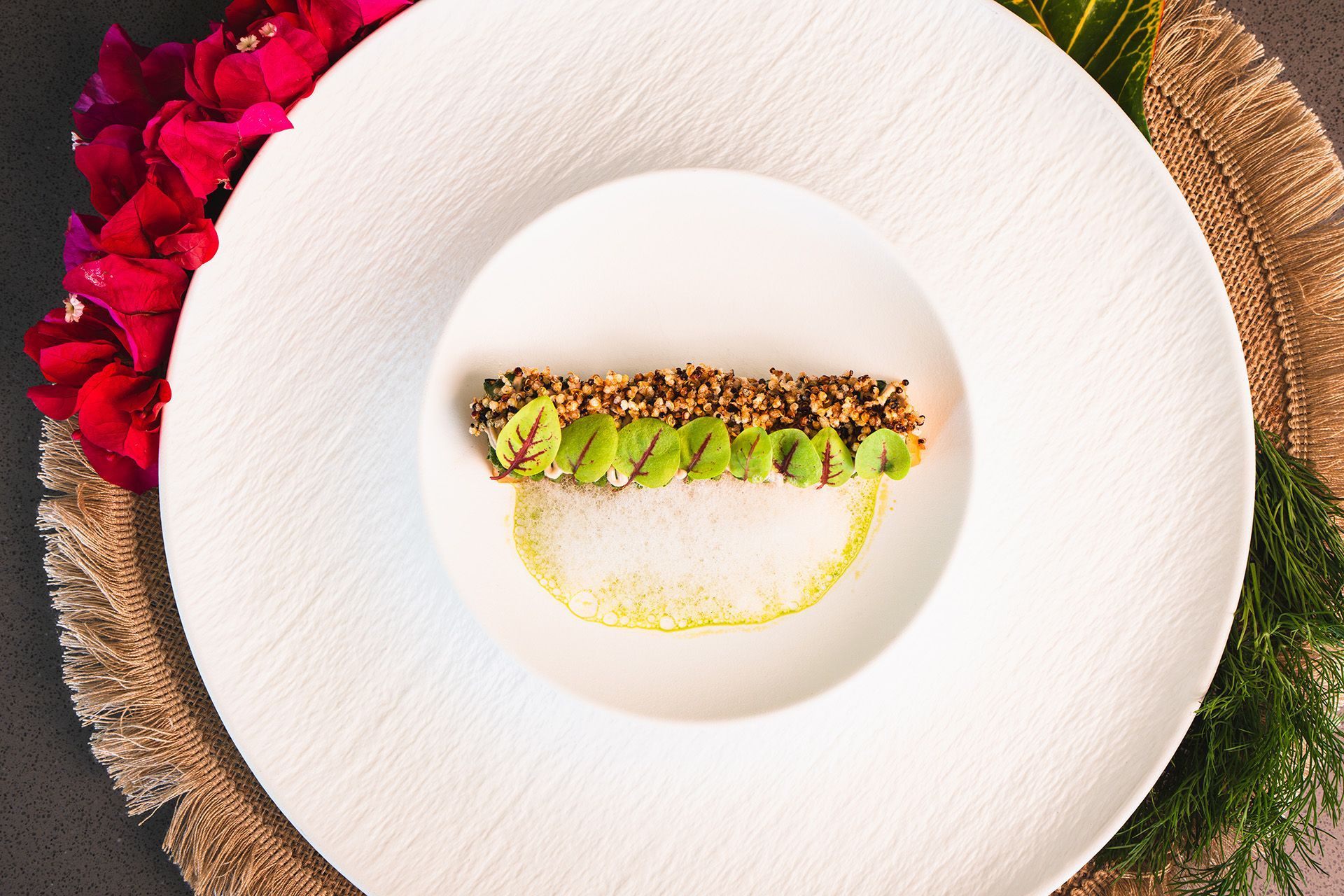 Elegant plated dish: Green and brown food atop a white plate with a yellow sauce, next to pink flowers.