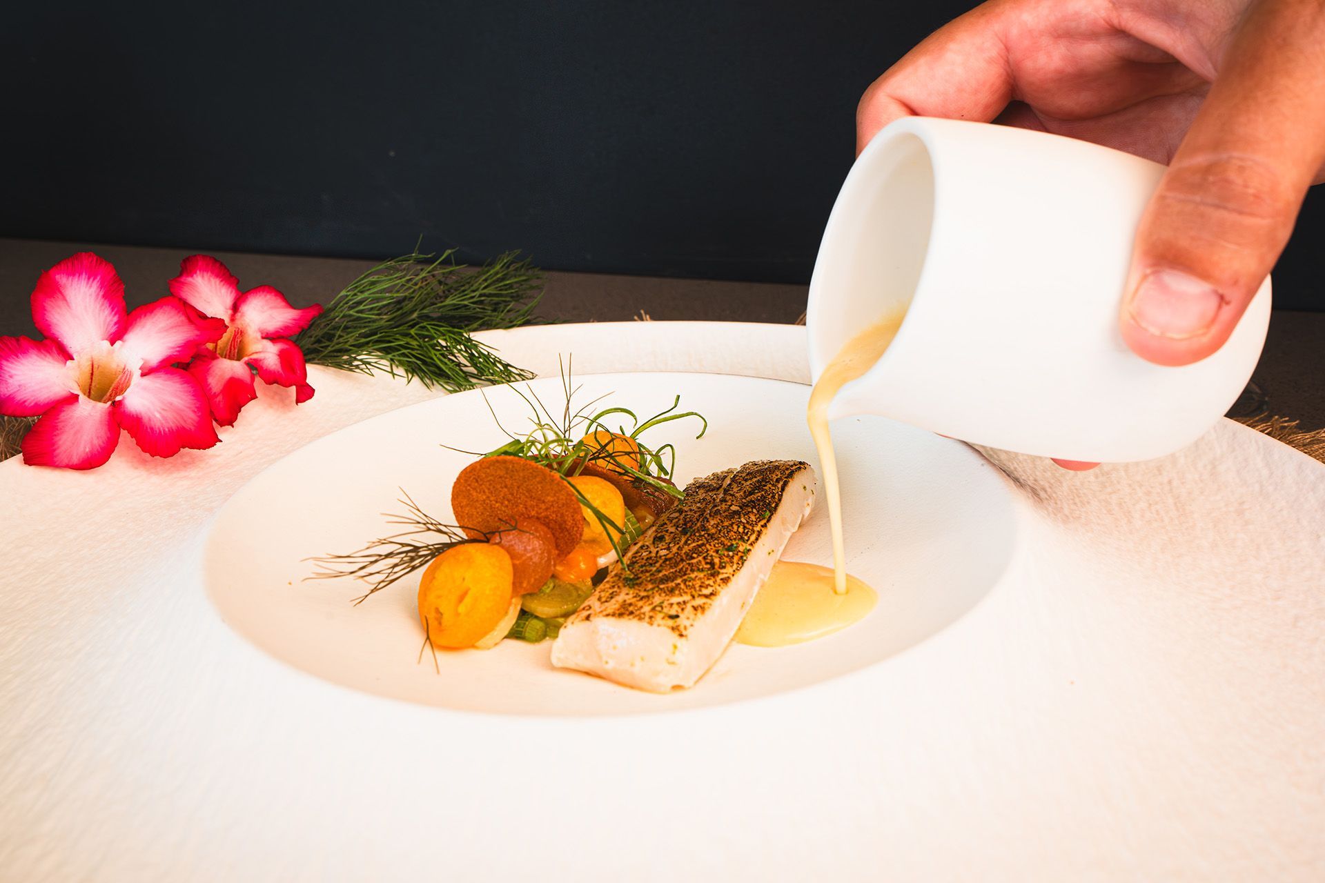 Hand pouring sauce over plated fish dish with vegetables; dark background.