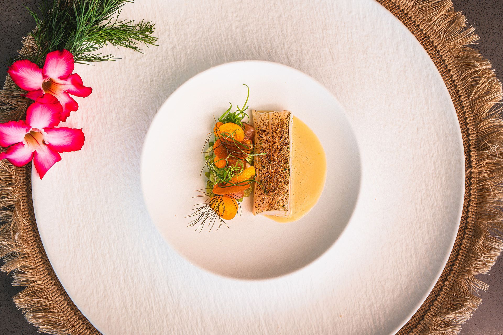 A plate of seared fish with sauce and garnish. Arranged on a textured white surface, with a flower on the side.