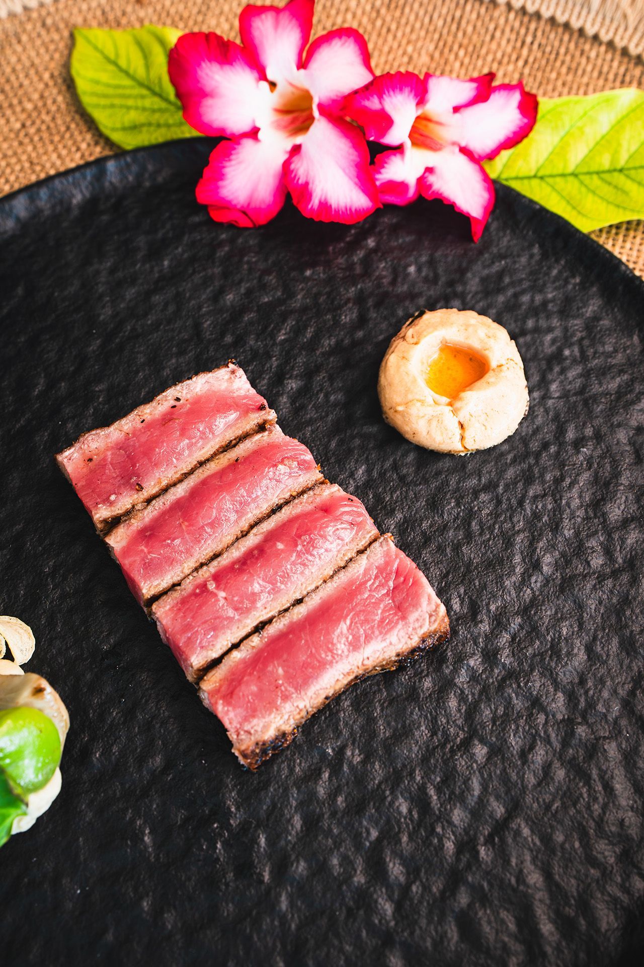 Sliced seared tuna on a black plate, with a cookie and pink flowers.