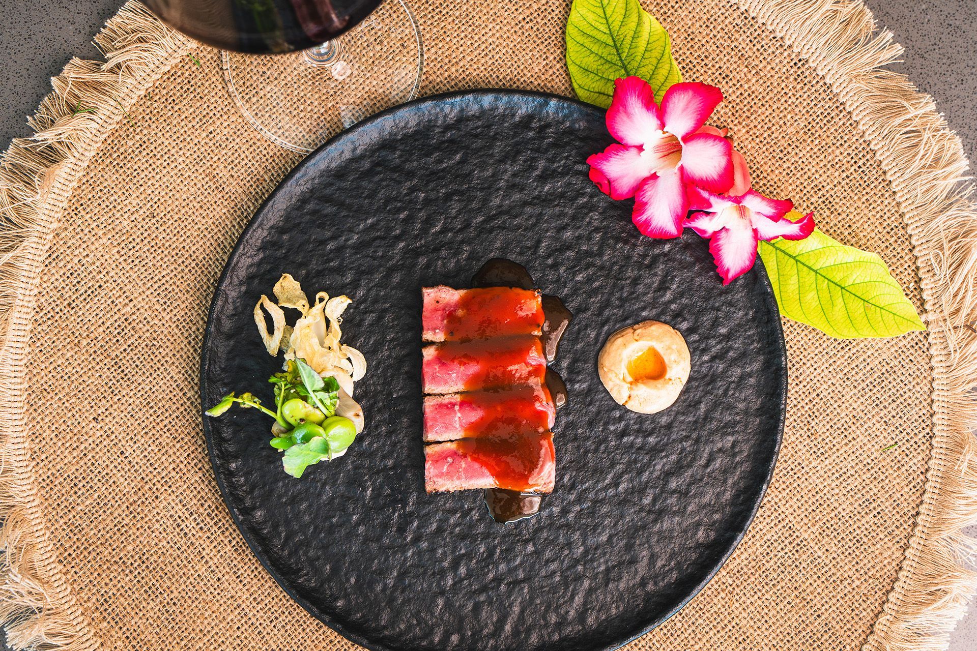 Seared tuna with sauce, served on a black plate with garnish and a flower, set on a tan mat.