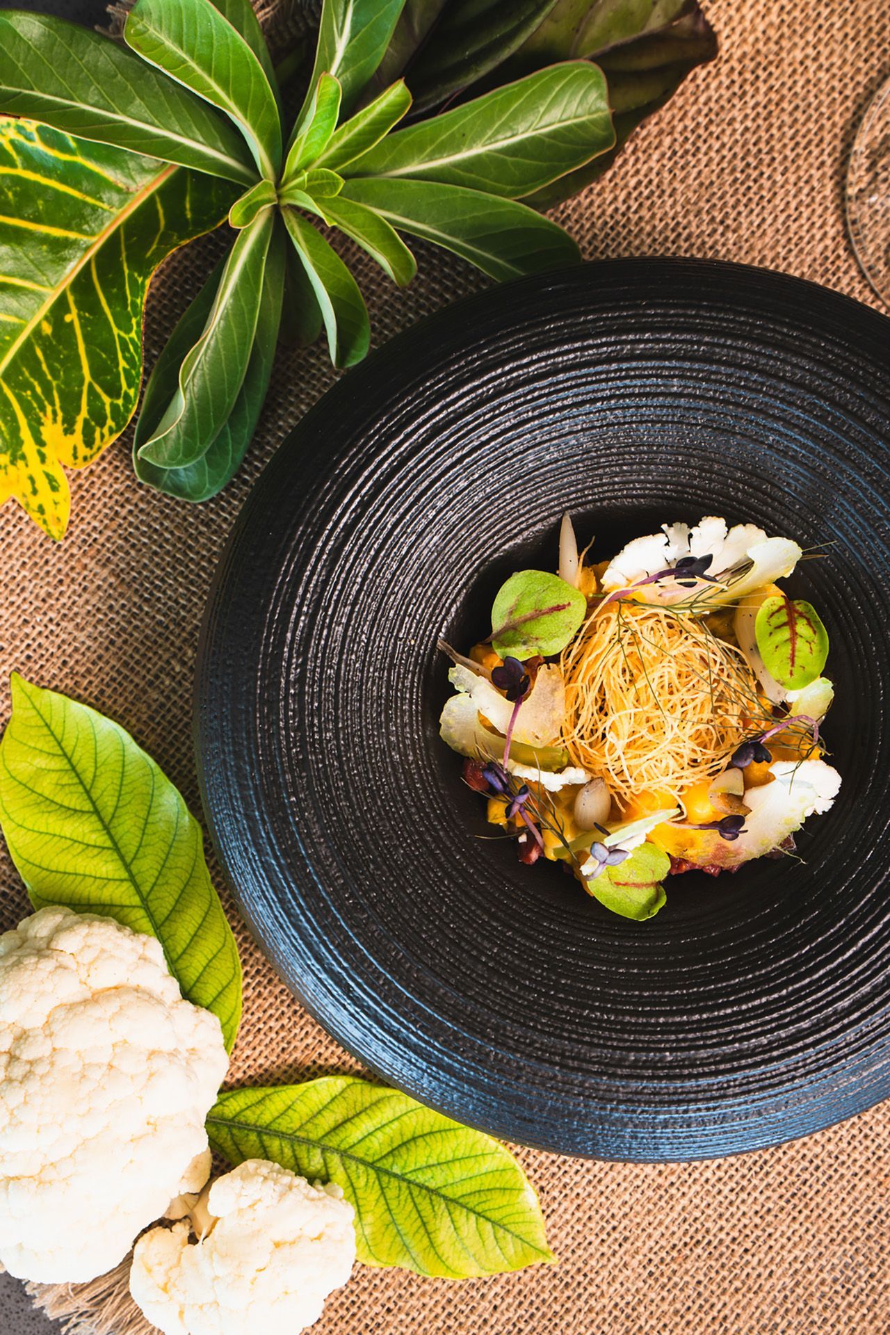 Black bowl with cauliflower salad and garnish, beside a whole cauliflower and leaves.