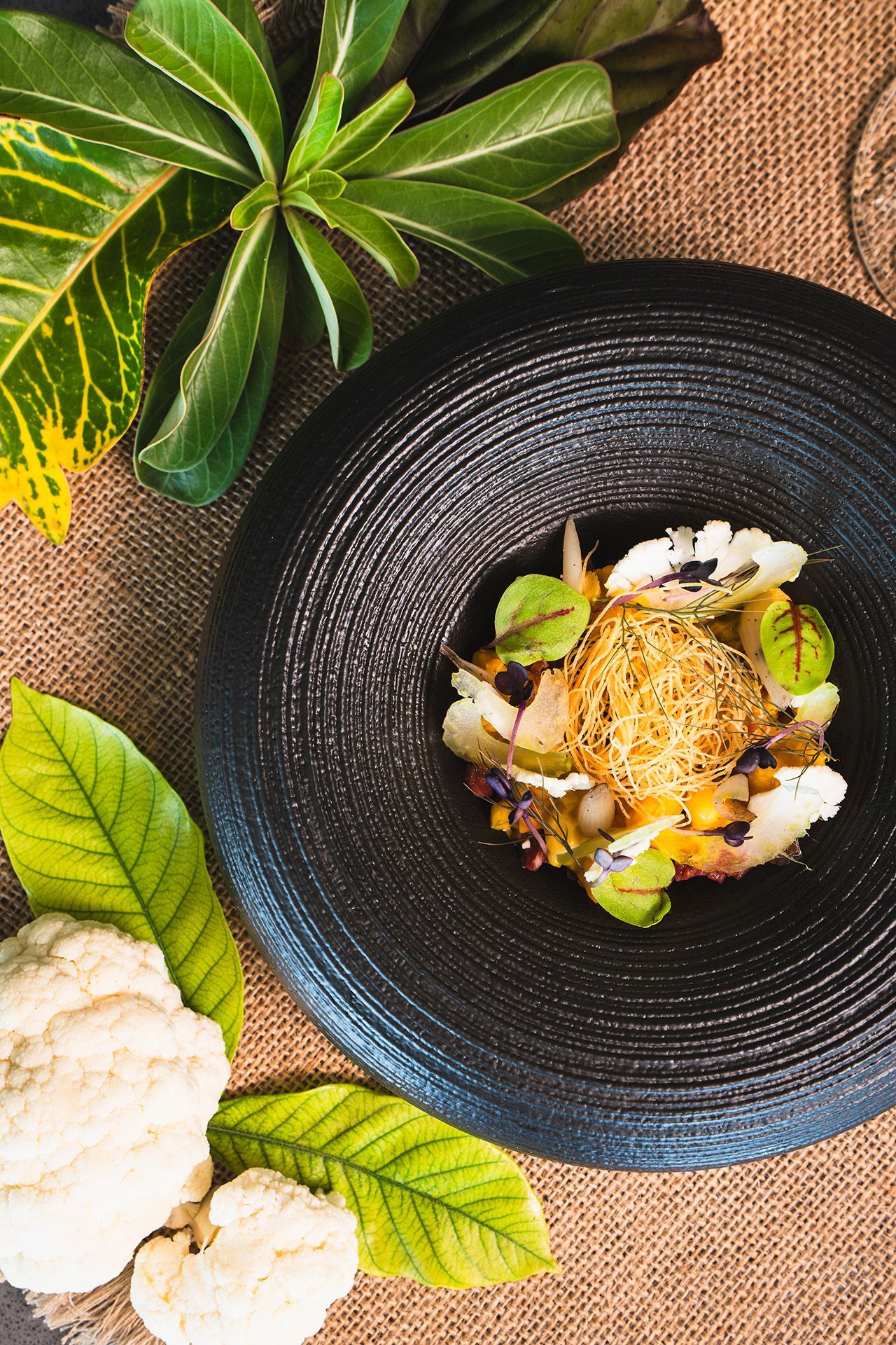 Gourmet cauliflower dish on a black plate with fresh leaves and cauliflower florets in a rustic setting.