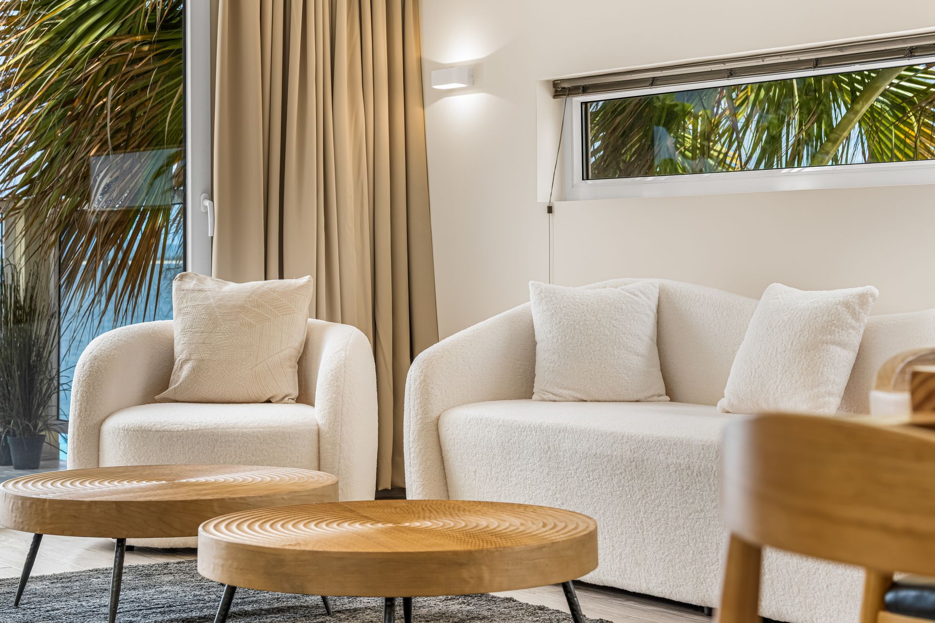 Bright living room with white sofa and chairs, round wooden tables, beige curtains, and a palm view through the window