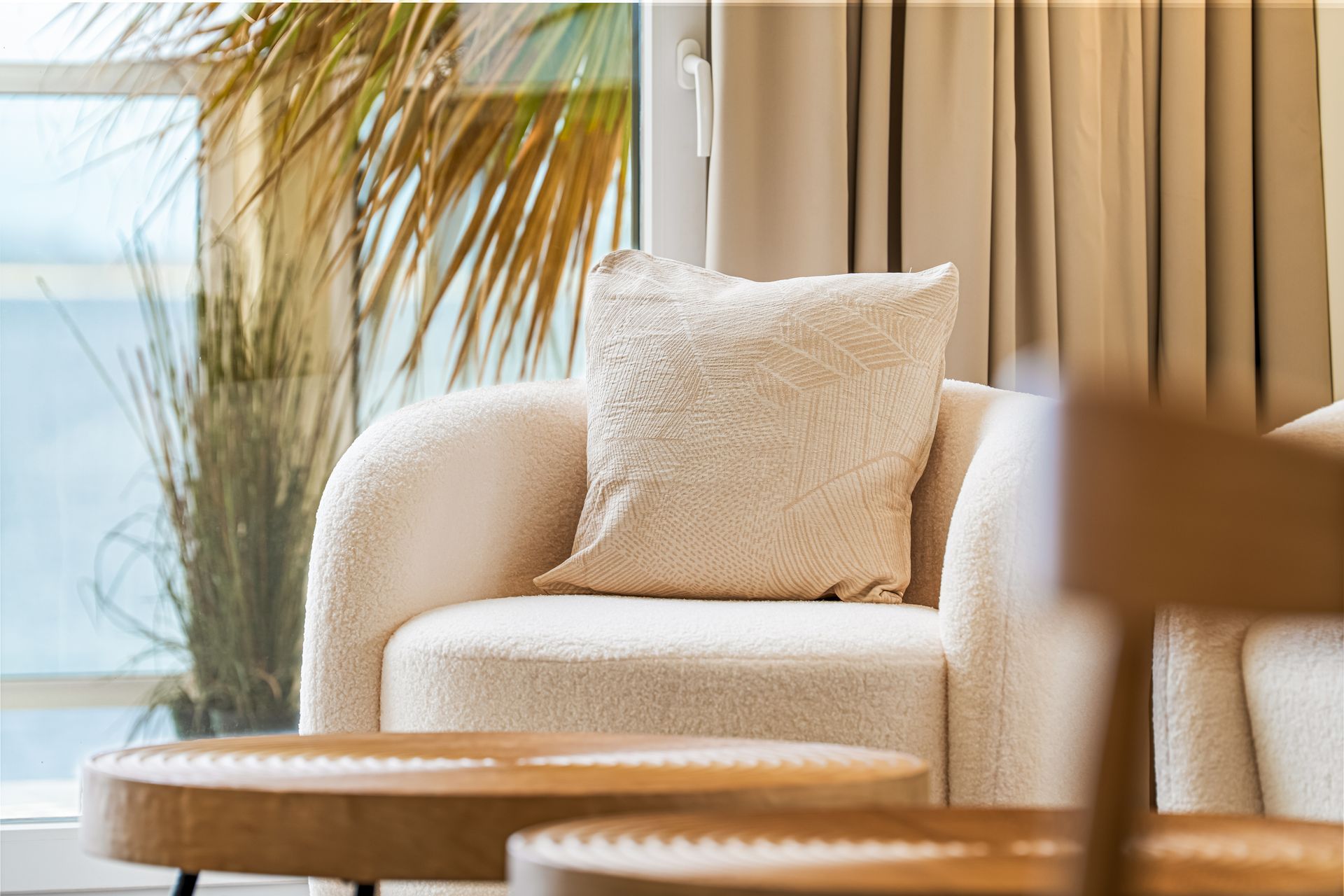 Cream armchair with pillow by a window, beside a potted palm and beige curtains in a bright room