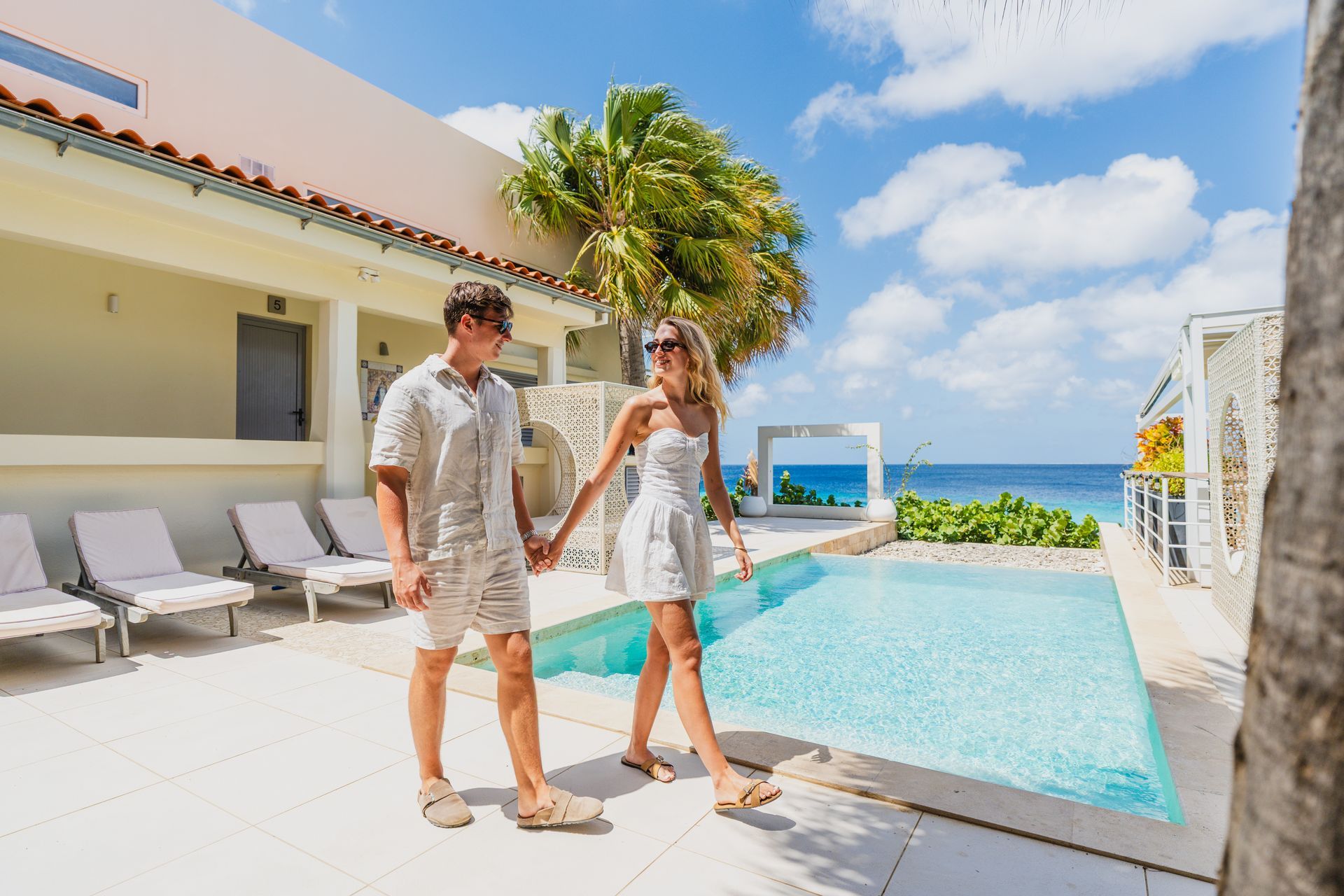 Couple holding hands by a pool at a beachfront resort with palm trees and blue ocean backdrop