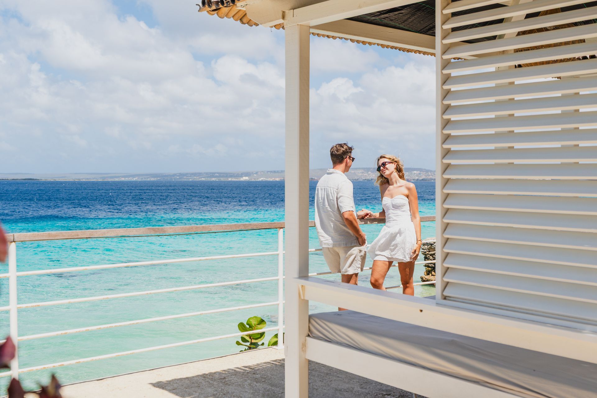 Two people relax on a balcony overlooking a bright turquoise ocean under a partly cloudy sky