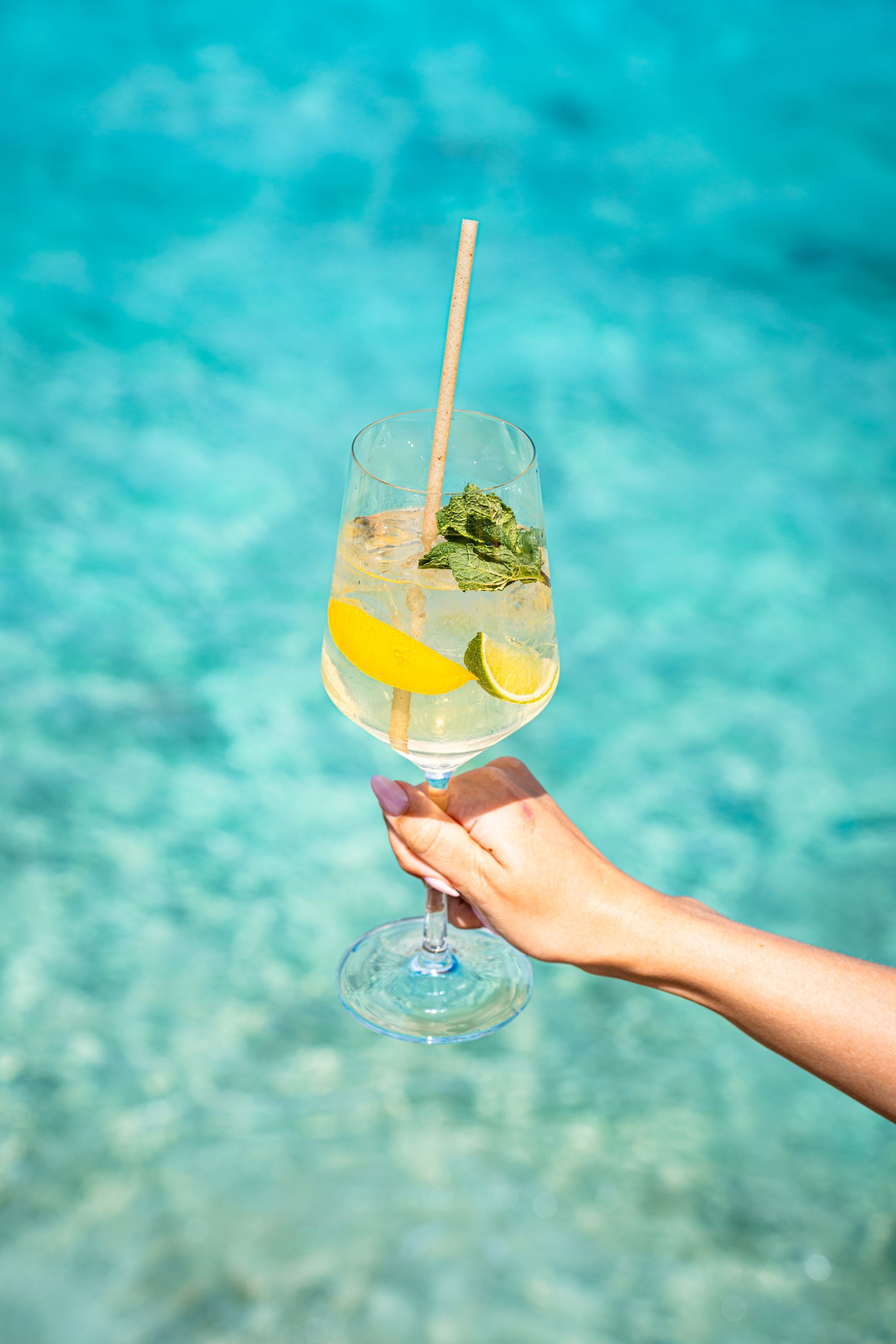 Hand holding a lemon mint cocktail with a straw against a turquoise sea background