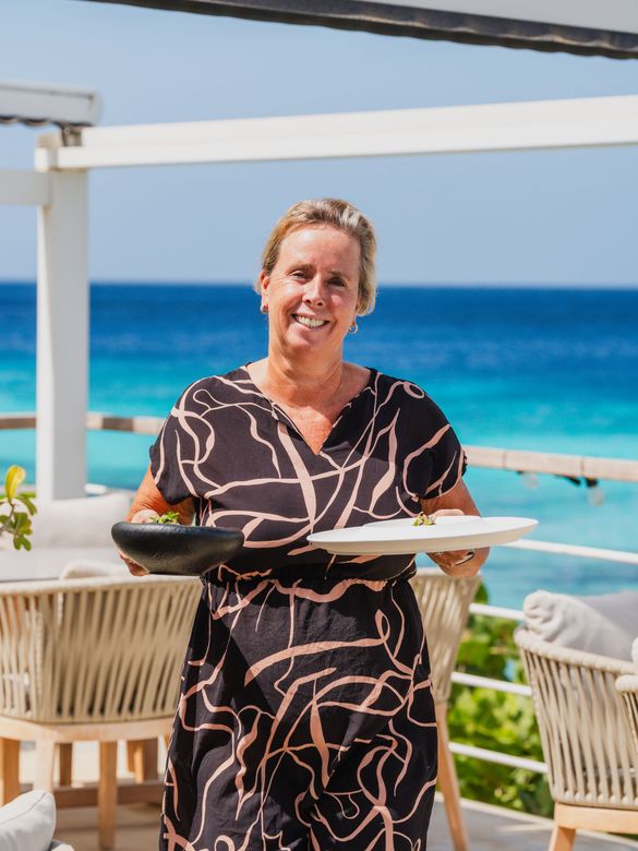 Smiling person carrying plates on a seaside patio with ocean in the background