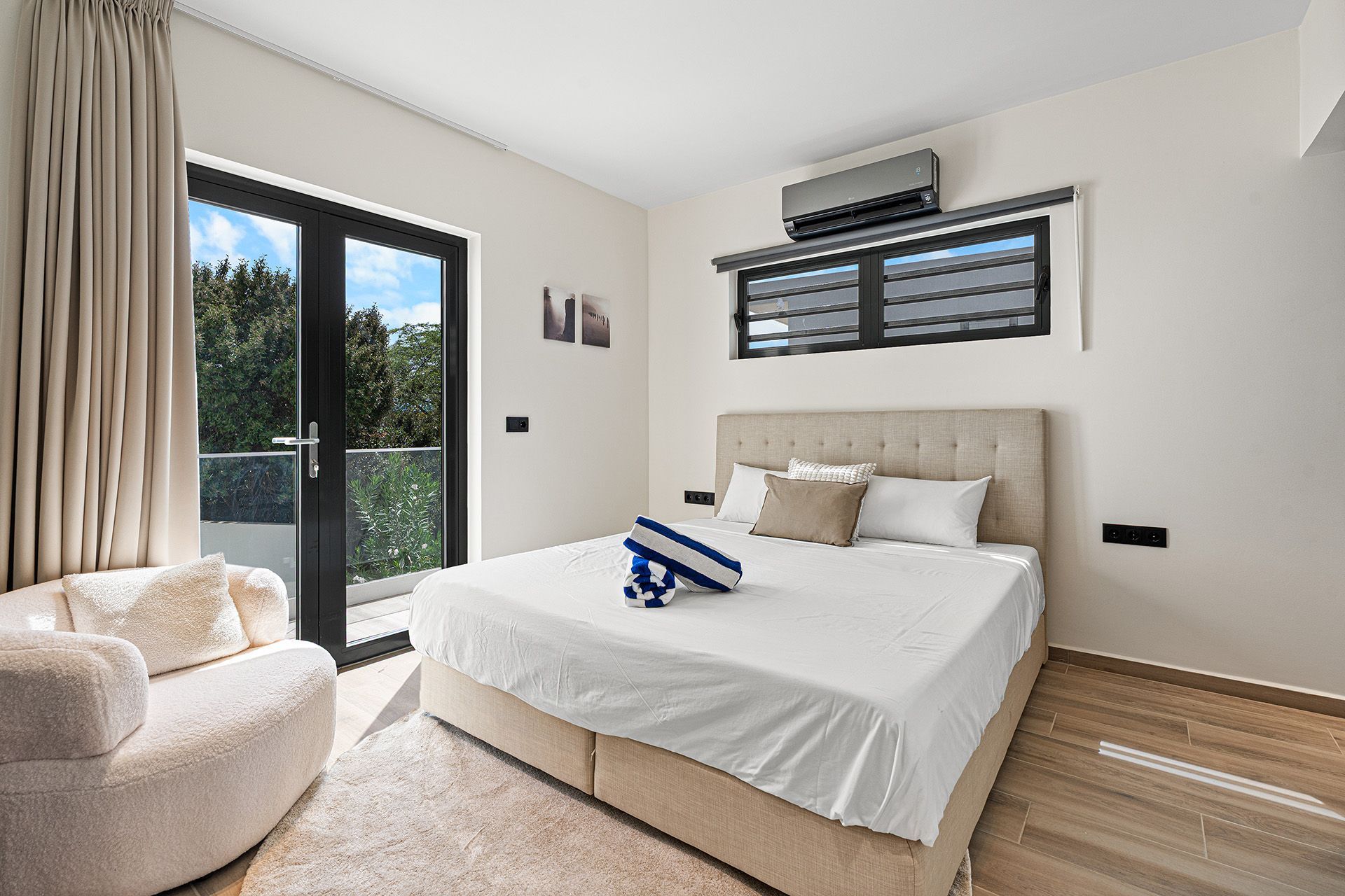 Bedroom with bed, balcony door, and armchair. Neutral tones with sunlight and outdoor view.
