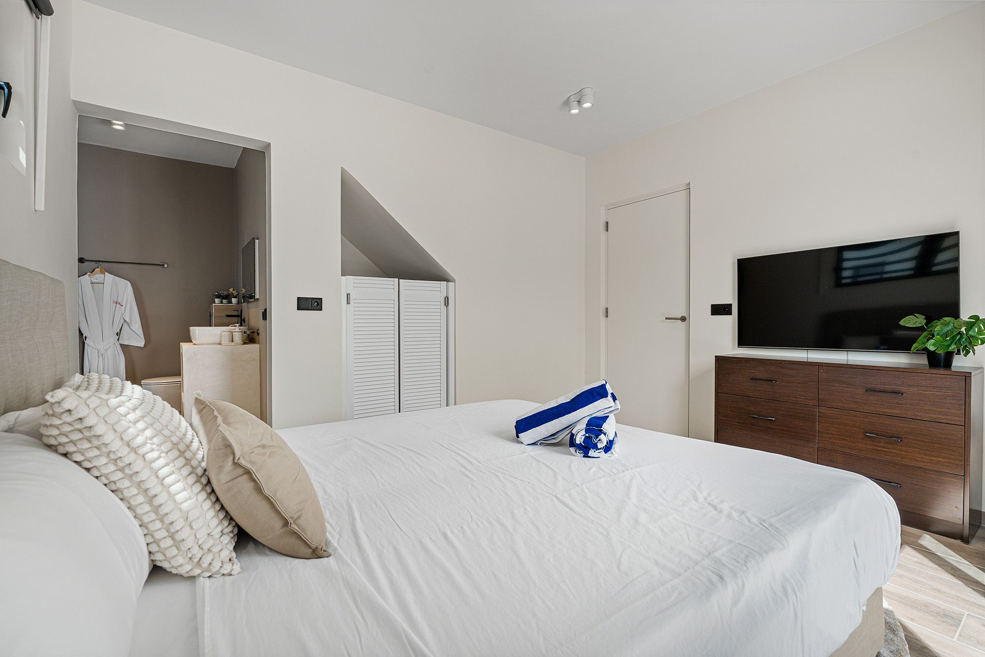 Bedroom with a large bed, TV on a dresser, and an adjacent bathroom. White and wood tones.