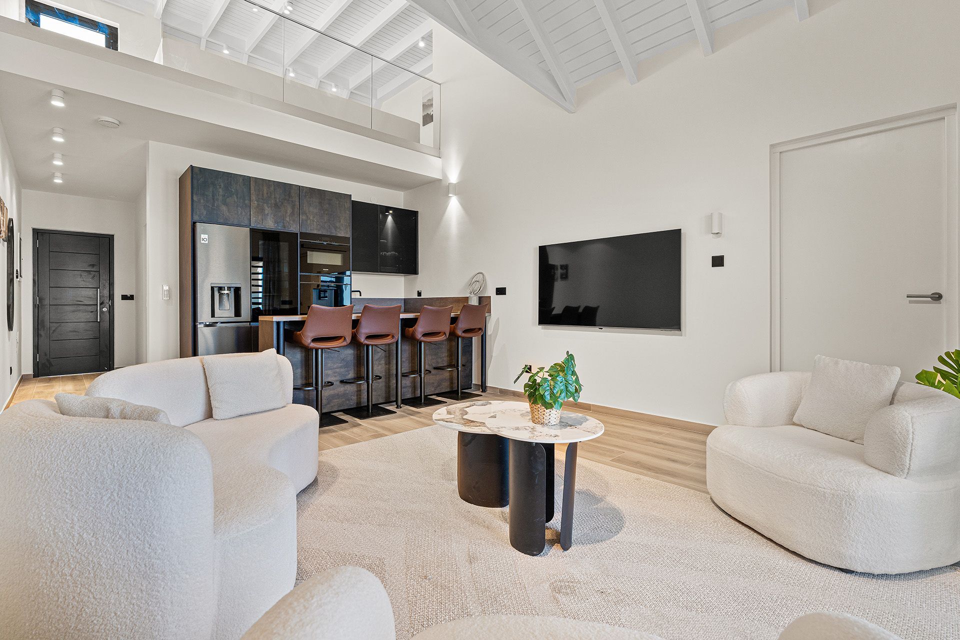 Modern living room with kitchen, white couches, bar stools, and TV.