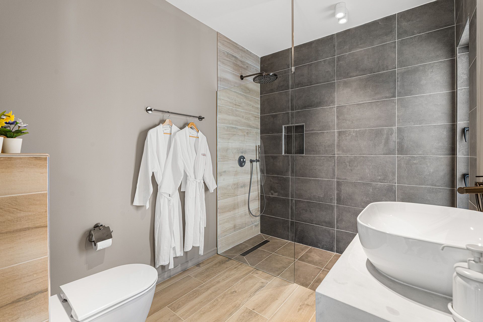 Modern bathroom with gray and light-colored tiled walls, shower, toilet, sink, and two white bathrobes hanging.