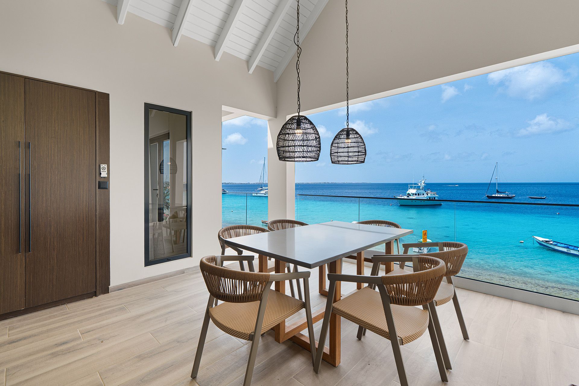 Dining room with square table, woven chairs, and ocean view through large windows.