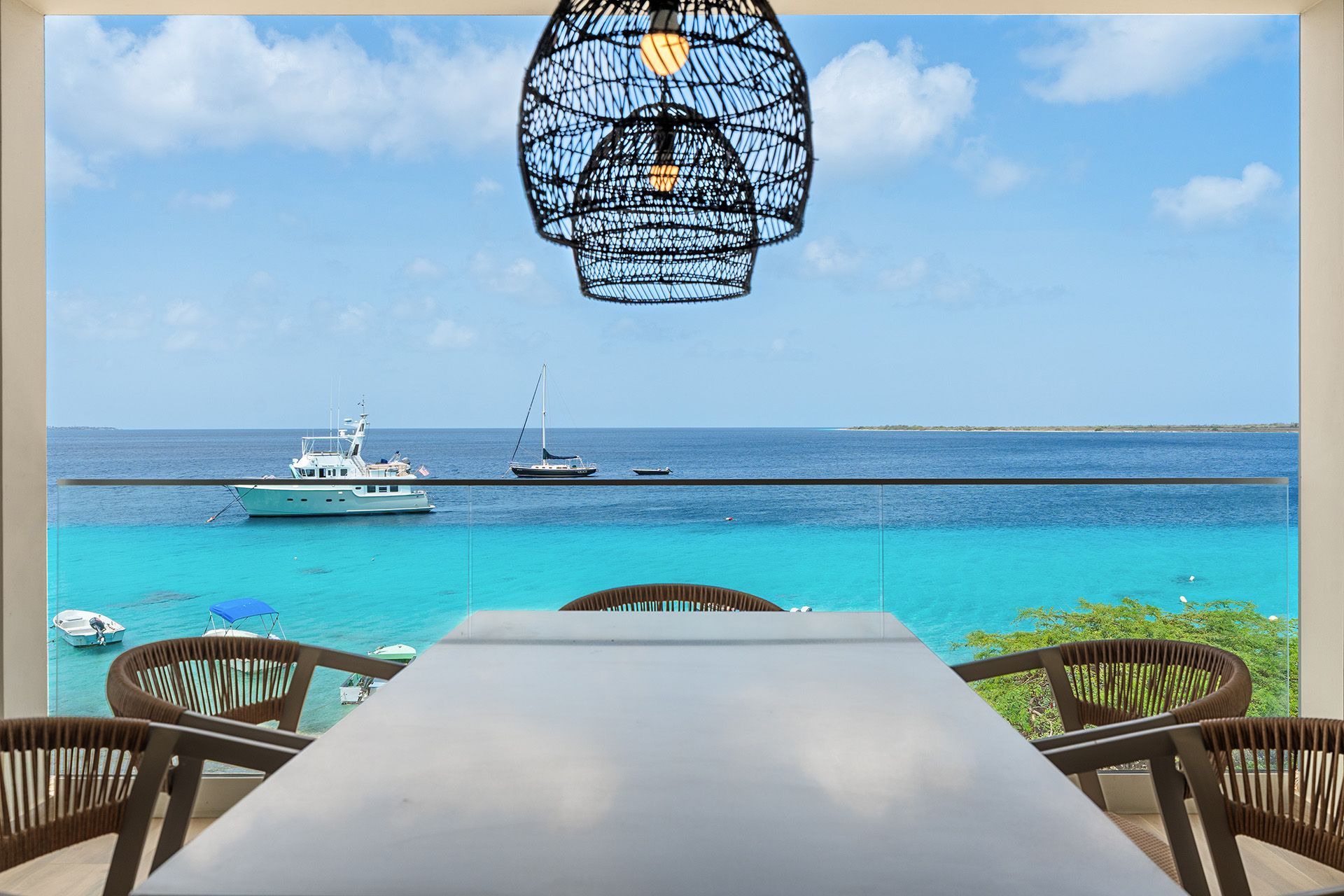 View of boats on turquoise water from a balcony with a white table and woven chairs, under a hanging lamp.