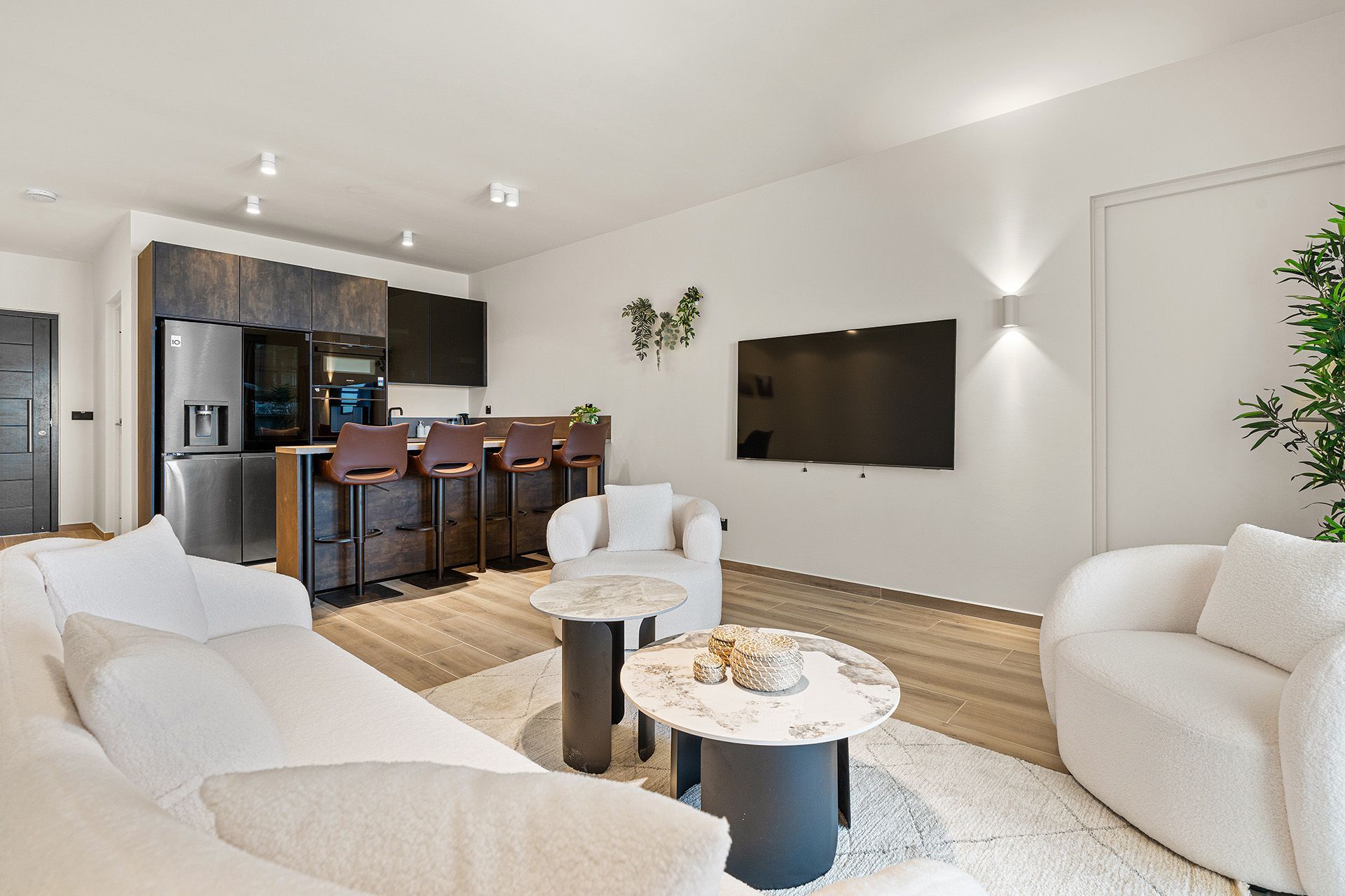 Modern living room with kitchen, white sofa, armchairs, two marble-topped tables, and mounted TV.