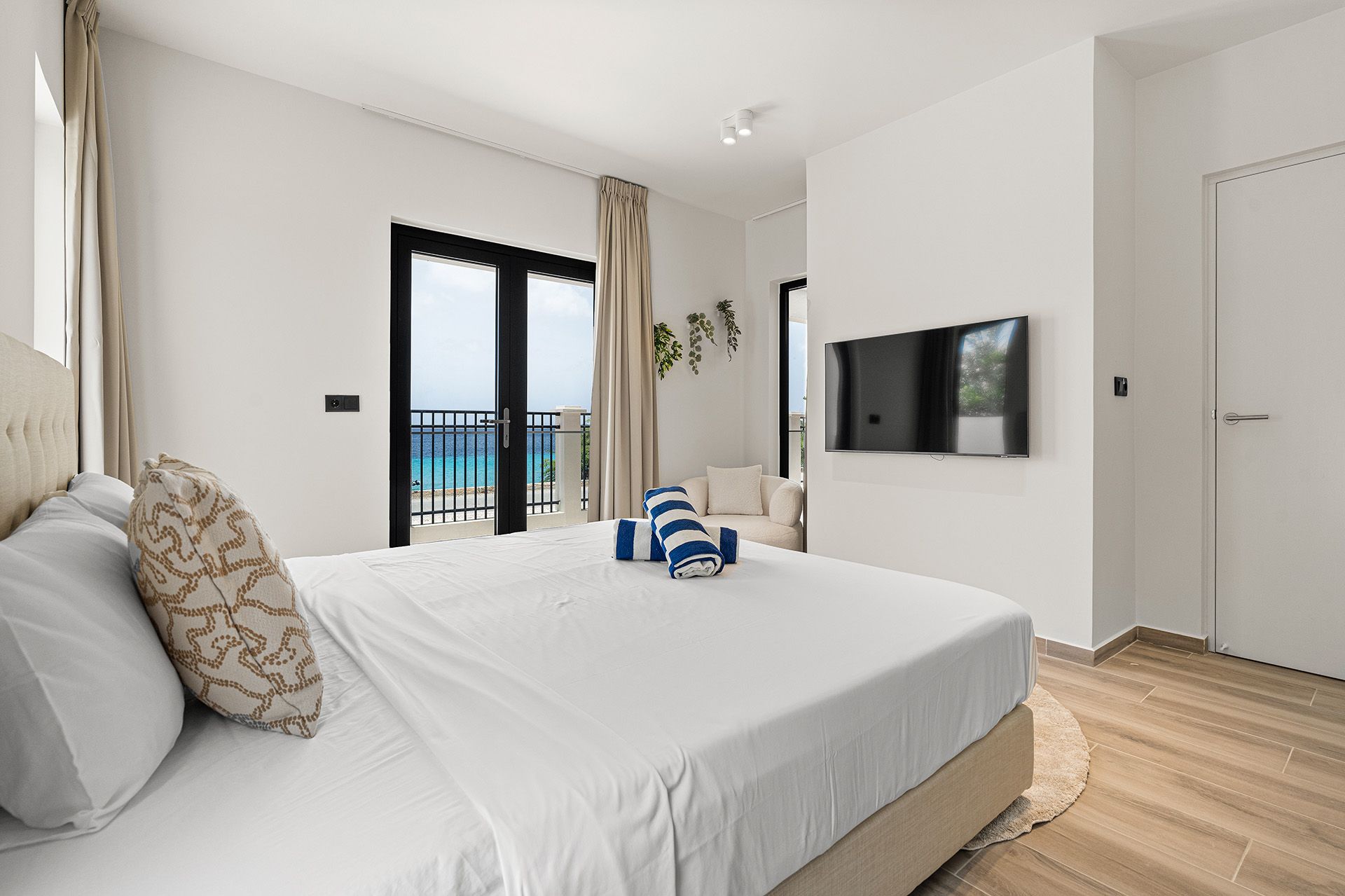 Bright bedroom with a king bed, TV, and balcony overlooking the ocean. White walls, light wood floors, and neutral tones.