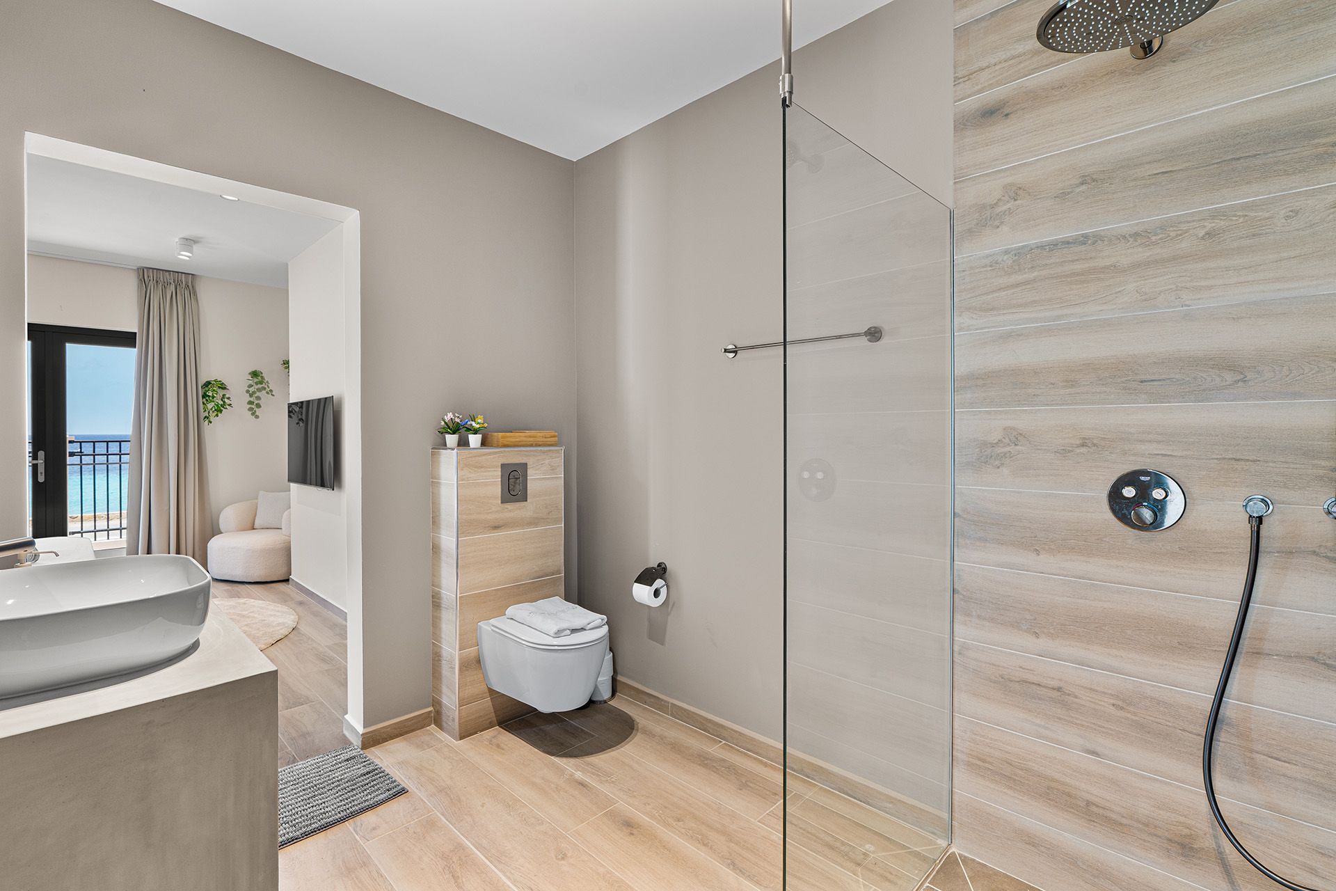 Bathroom with glass shower, light wood floors, and view of the sea from an open doorway.