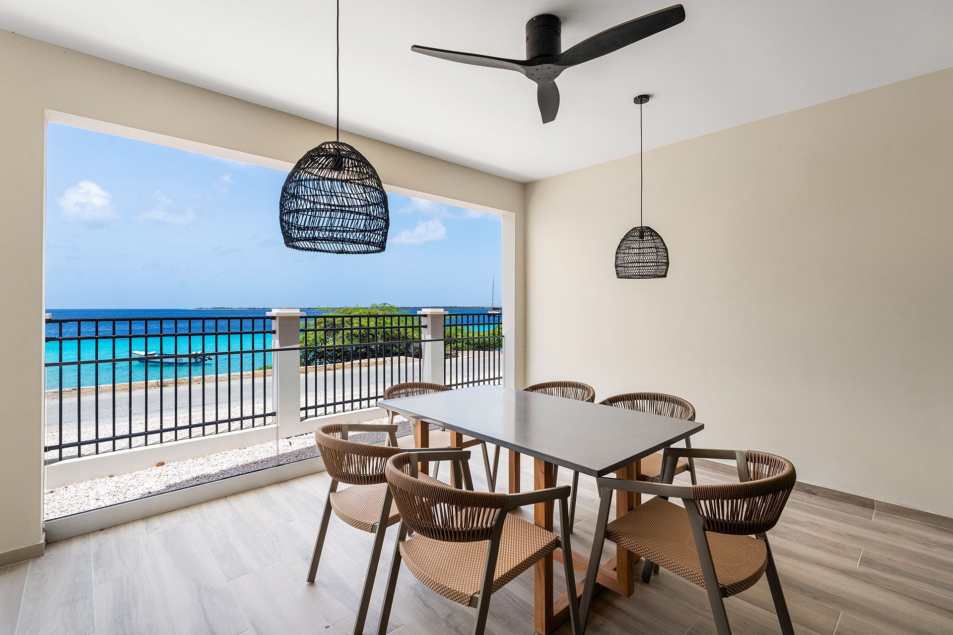 Dining room with a balcony overlooking a blue ocean. Table and chairs with hanging pendant lights and a ceiling fan.