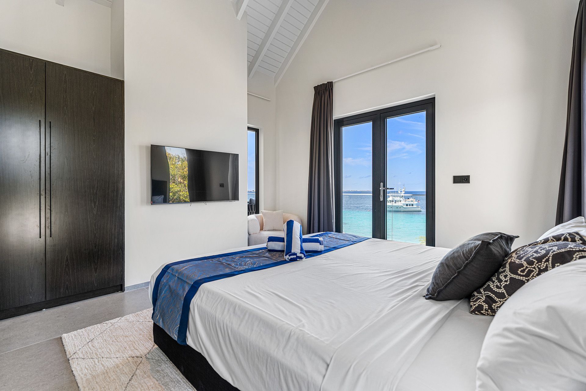 Bedroom with a bed, TV, and view of the ocean through open doors. Features a dark wardrobe and white walls.