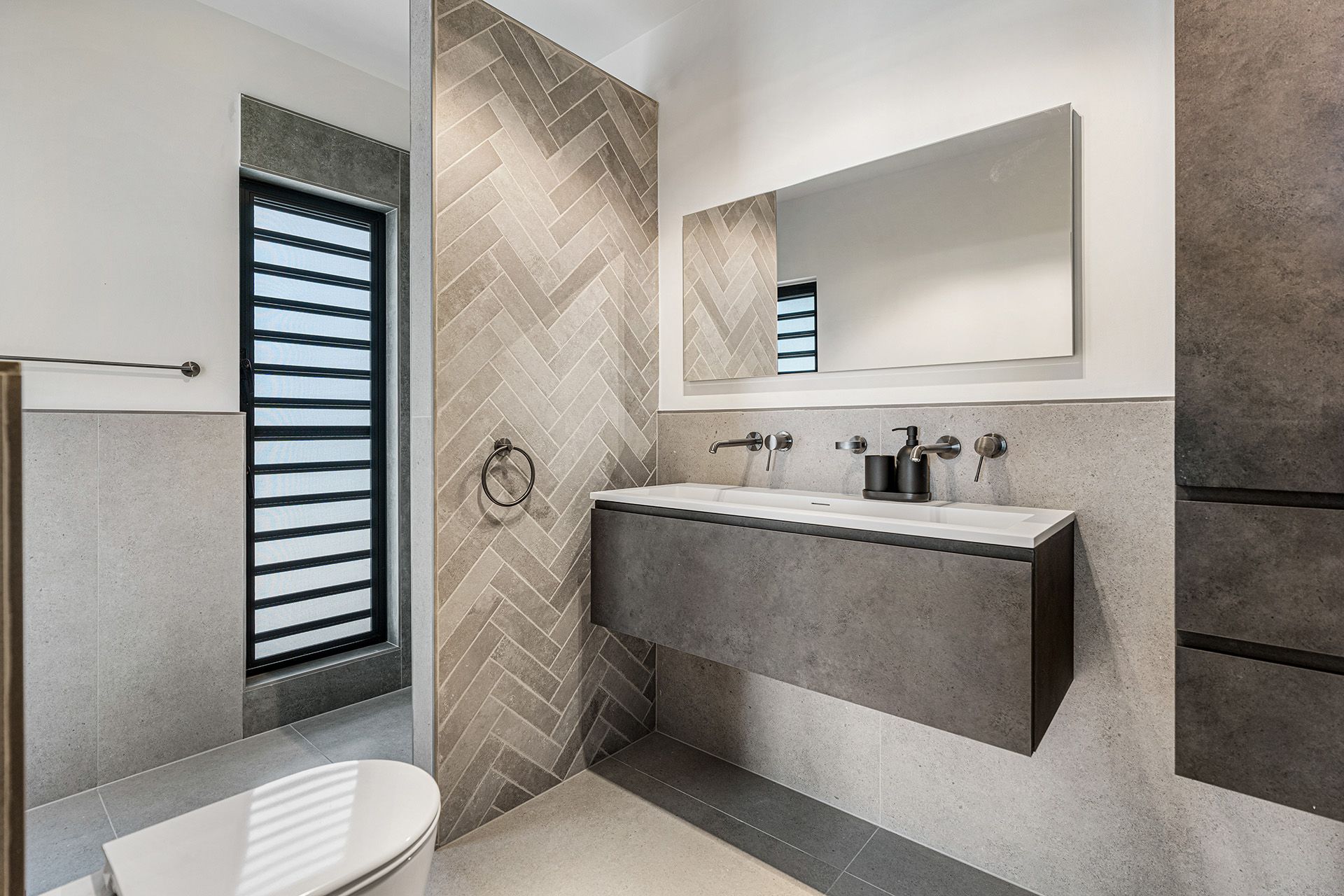 Modern bathroom with floating gray vanity, herringbone tile accent wall, and large mirror.