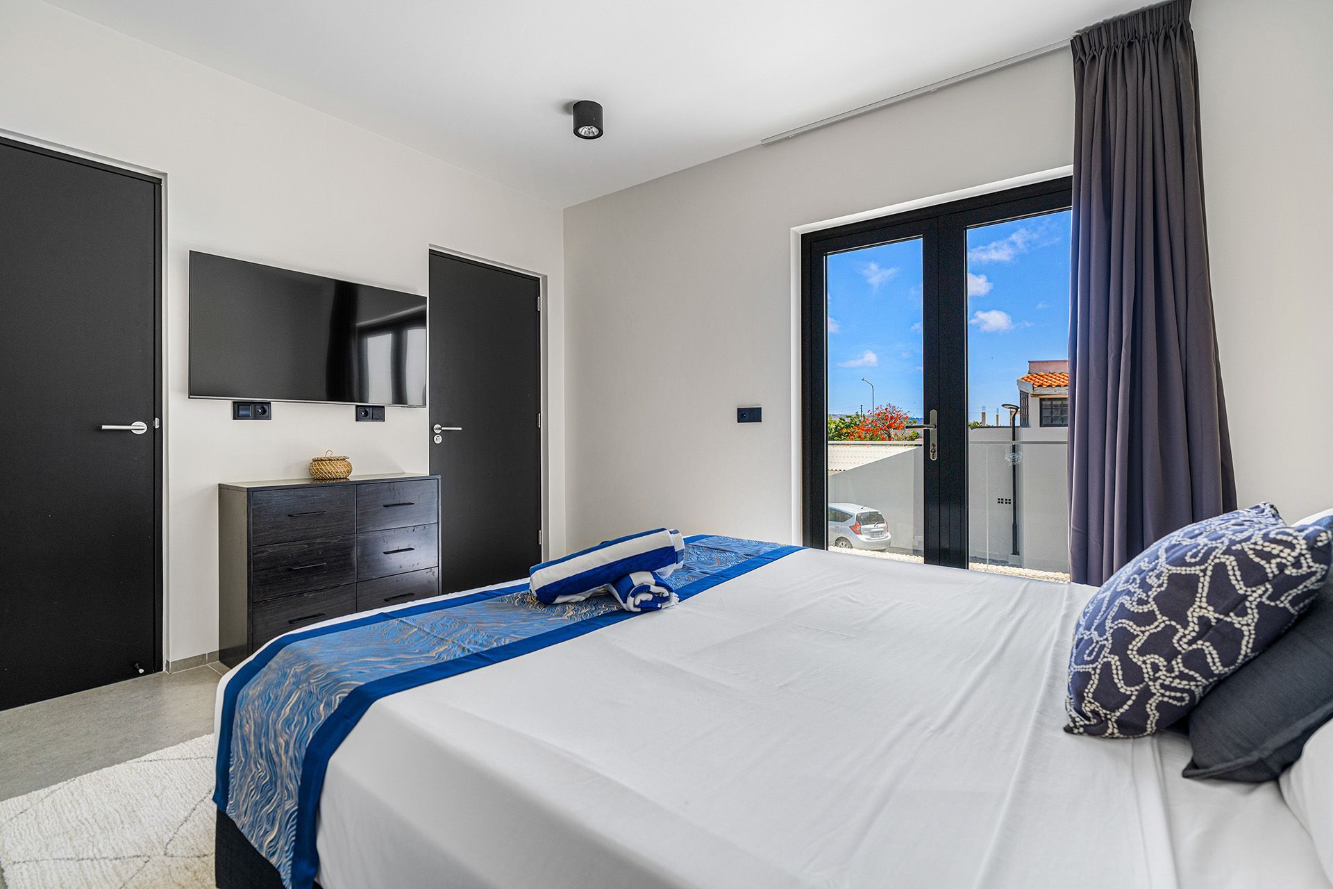Bedroom with a bed, dresser, doors, TV, and a door to a balcony with a blue sky.
