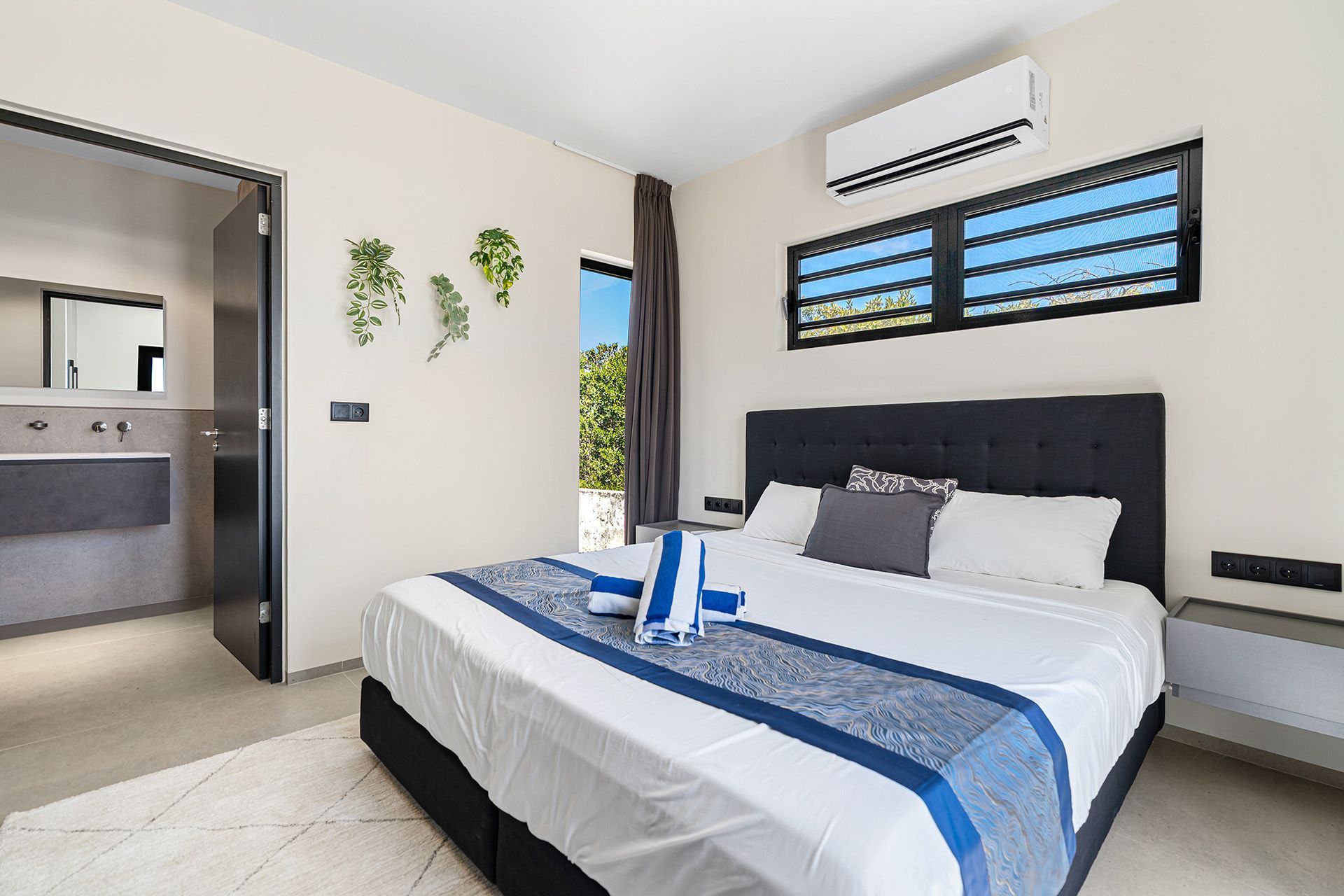 Bedroom with king bed, blue accents, bathroom doorway, and greenery outside window.