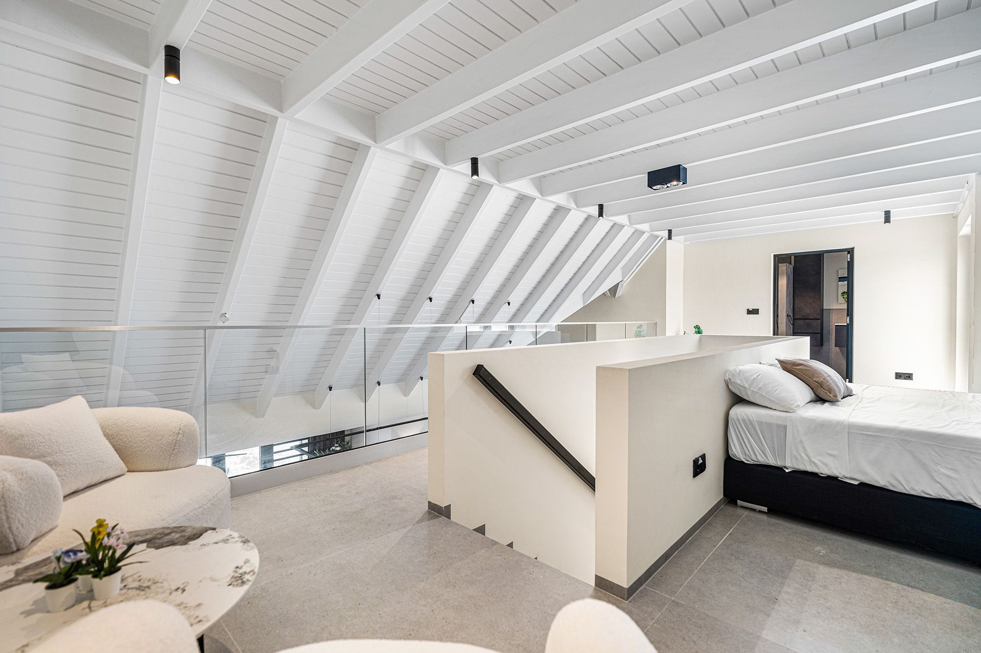 Modern loft interior with a bedroom, living area, and sloped white ceiling with exposed beams.