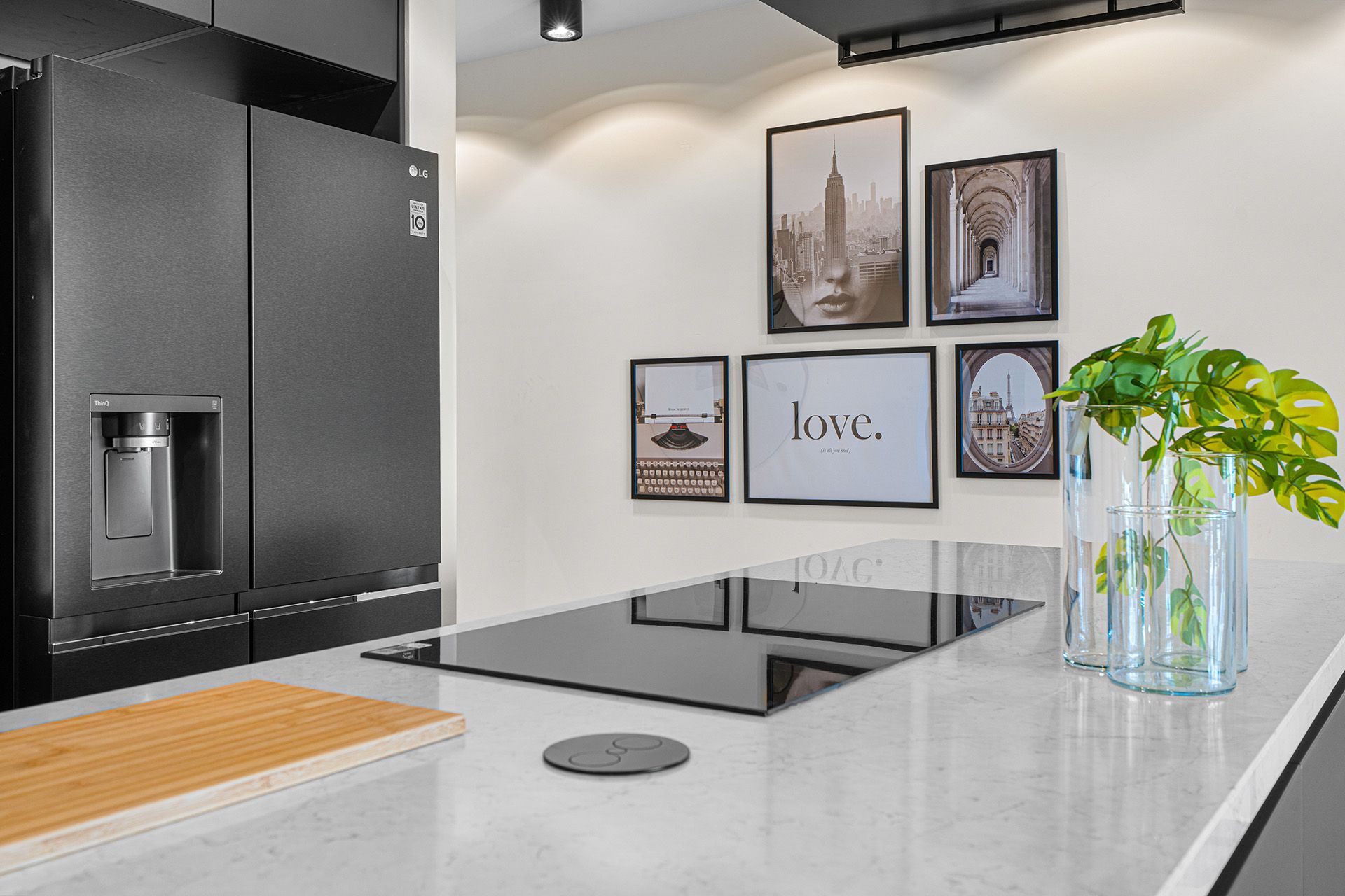 Black kitchen with a fridge, island with cooktop, framed art, and vase of green plants.