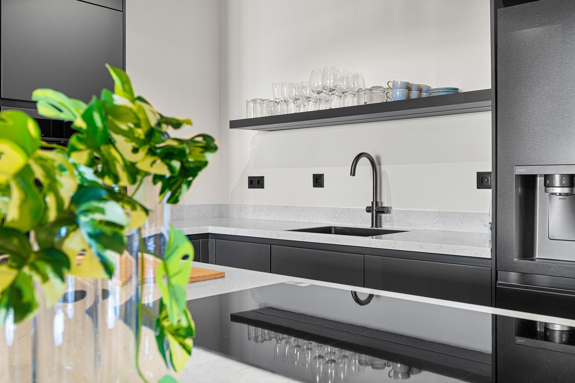 Modern kitchen with black cabinets, white countertops, and a stainless steel faucet. A plant sits in the foreground.