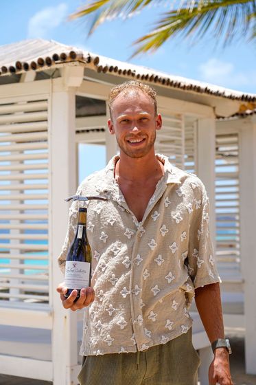 Person standing outdoors holding a bottle, wearing a beige patterned shirt in front of a white beach house.