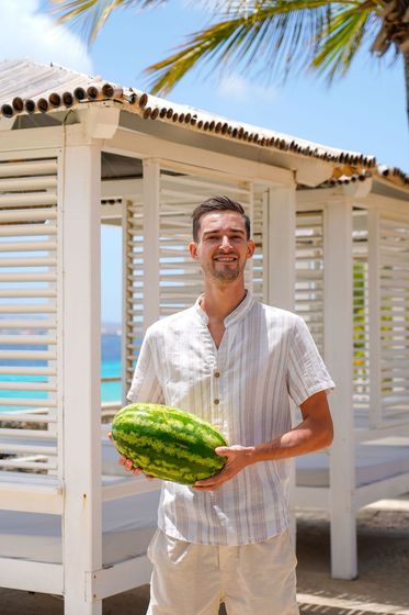 Man holding a watermelon in a white beach cabana by the ocean
