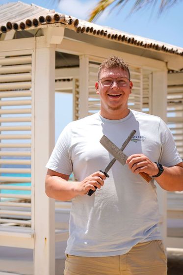 Man smiling outdoors, holding two crossed metal tools in front of a white beach pavilion.