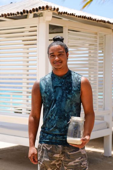 Man holding a glass jar outdoors beside a white louvered structure, wearing a sleeveless blue top and camo shorts.