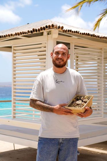 Man holding a tray of food beside a white beach cabana with turquoise water in the background