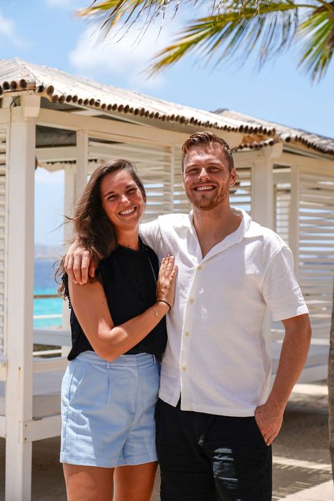 Two people smiling outdoors by a white beach house, standing arm-in-arm in bright sunlight