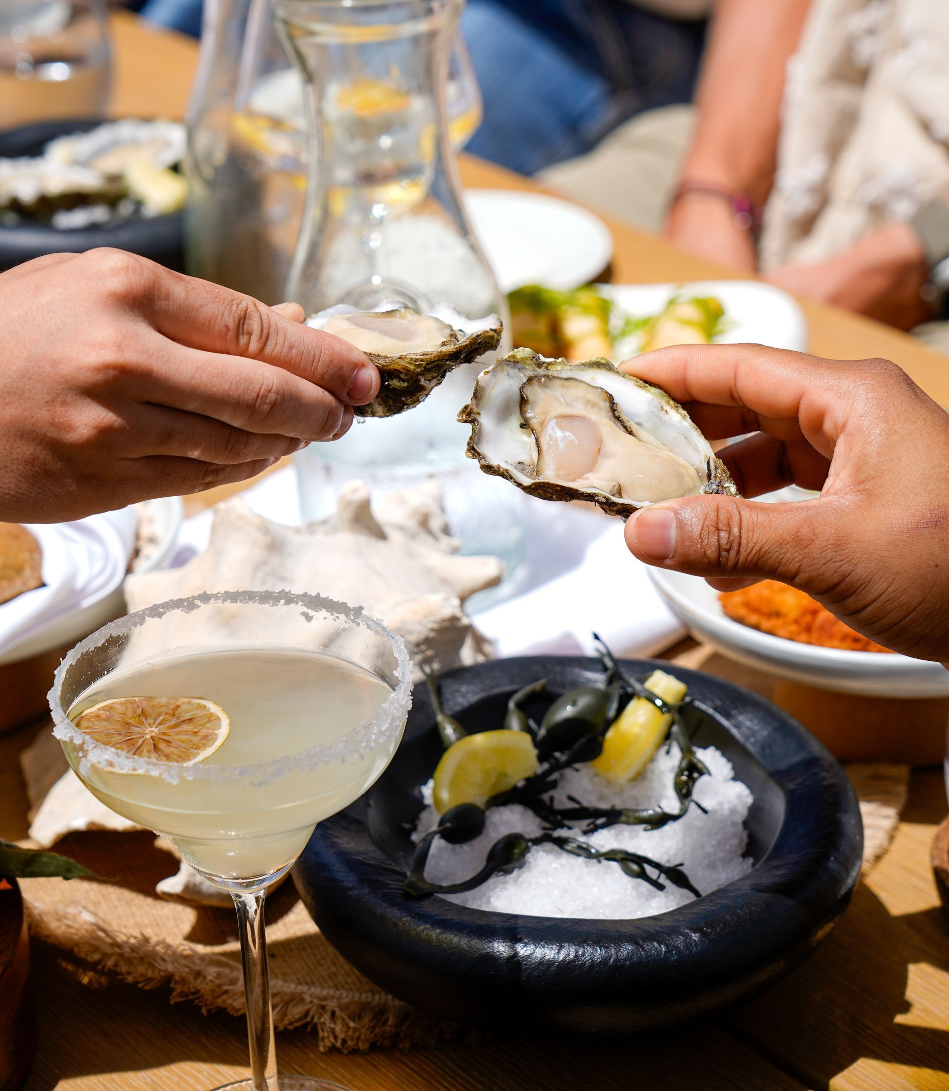 Hands opening an oyster at a seafood table with lemon, wine, and plates of shellfish