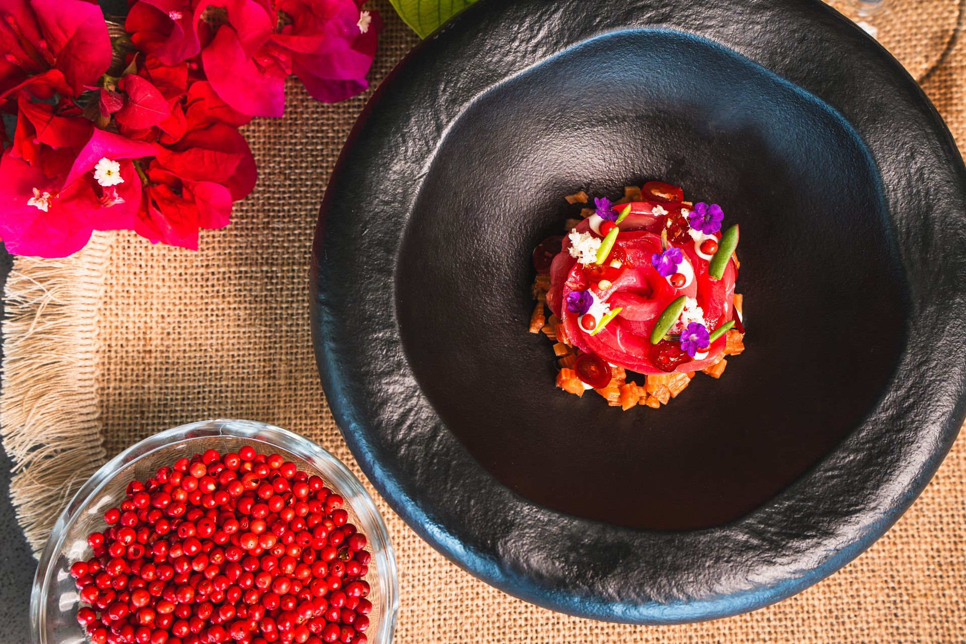 Gourmet dessert in a dark bowl, topped with red sauce, flowers, and peppercorns. A bowl of peppercorn, pink flowers