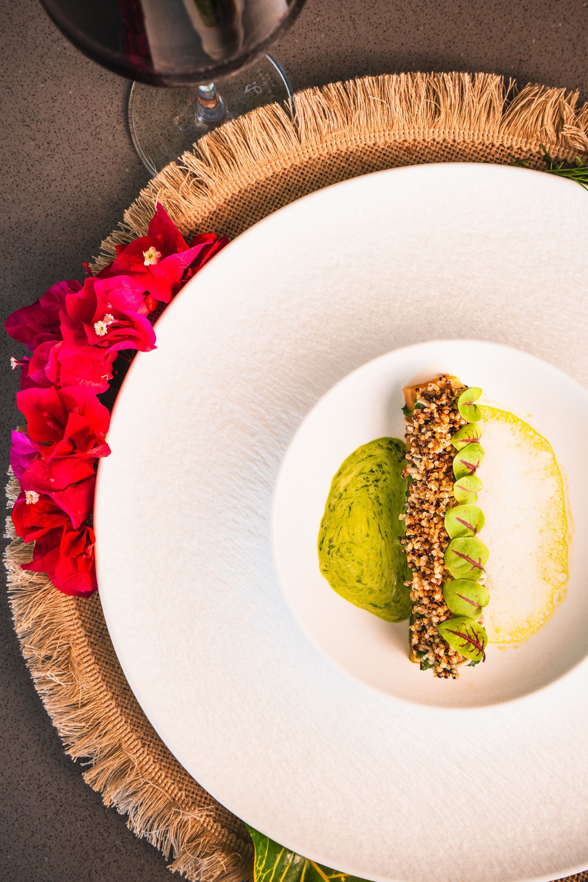 Overhead view of a plated dessert with green and white components, red flowers, and a glass of red wine.