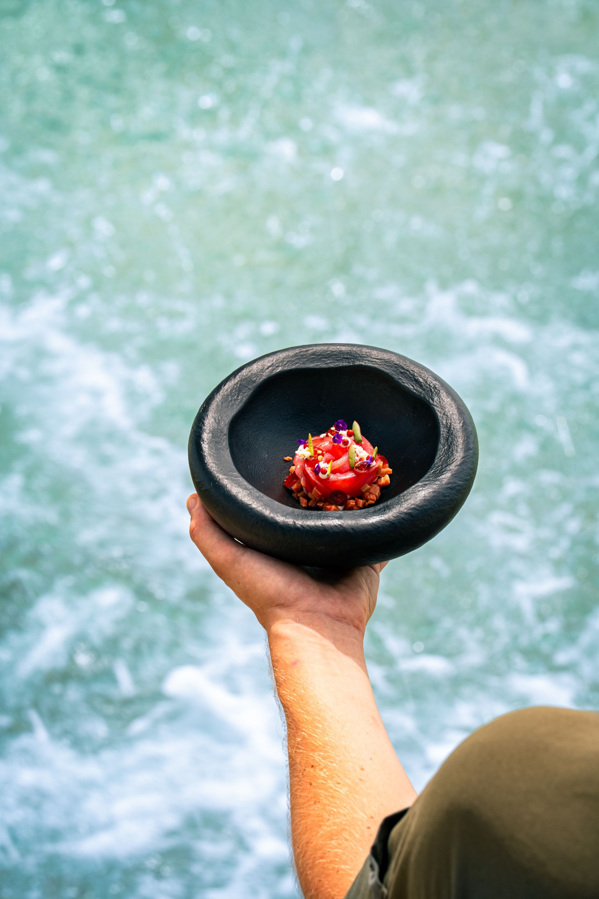 Hand holding a black bowl with a red dish, blurred water background.