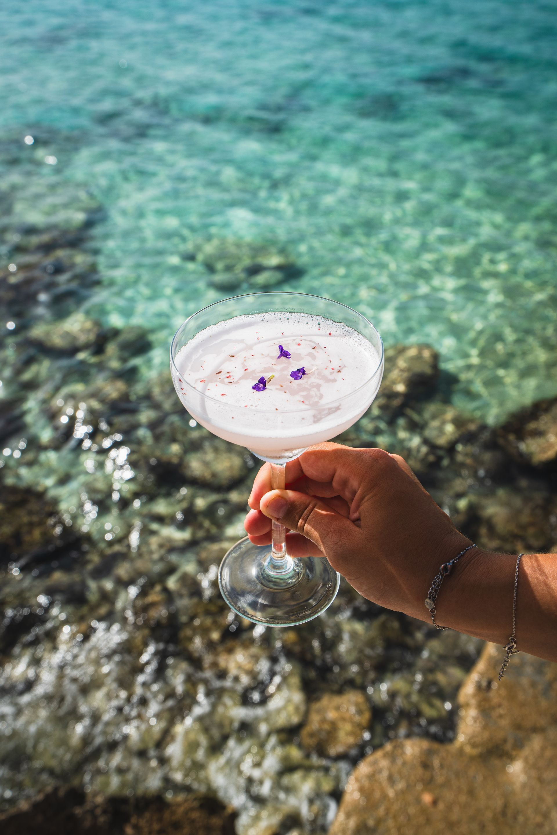 Hand holding a cocktail garnished with purple flowers, overlooking clear turquoise water.