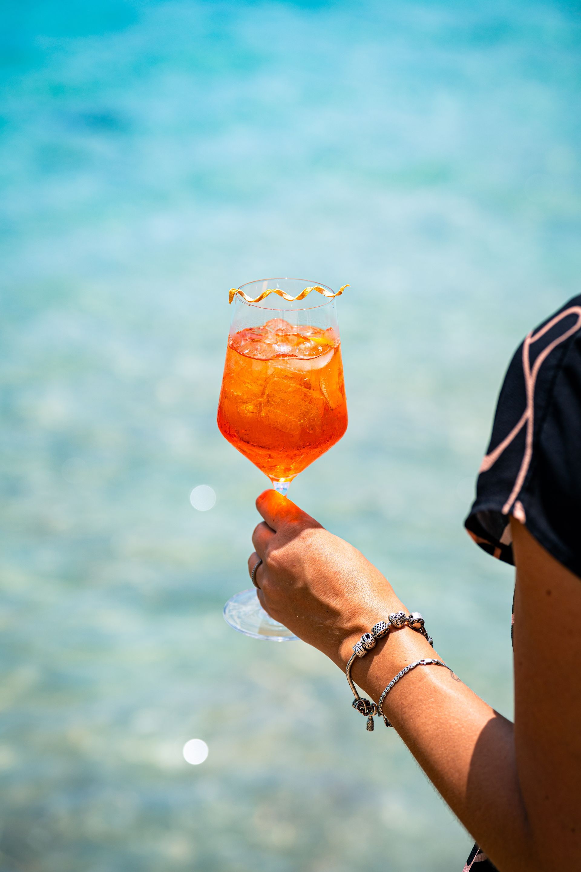 Person holding an orange cocktail in a stemmed glass, turquoise ocean in background.